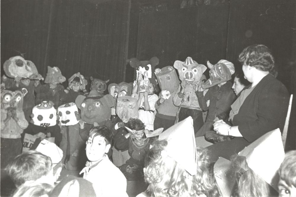 A group of children wearing large, handmade animal masks stand on stage, while two adults look on. The scene appears to be part of a school play or performance. The photo is in black and white.