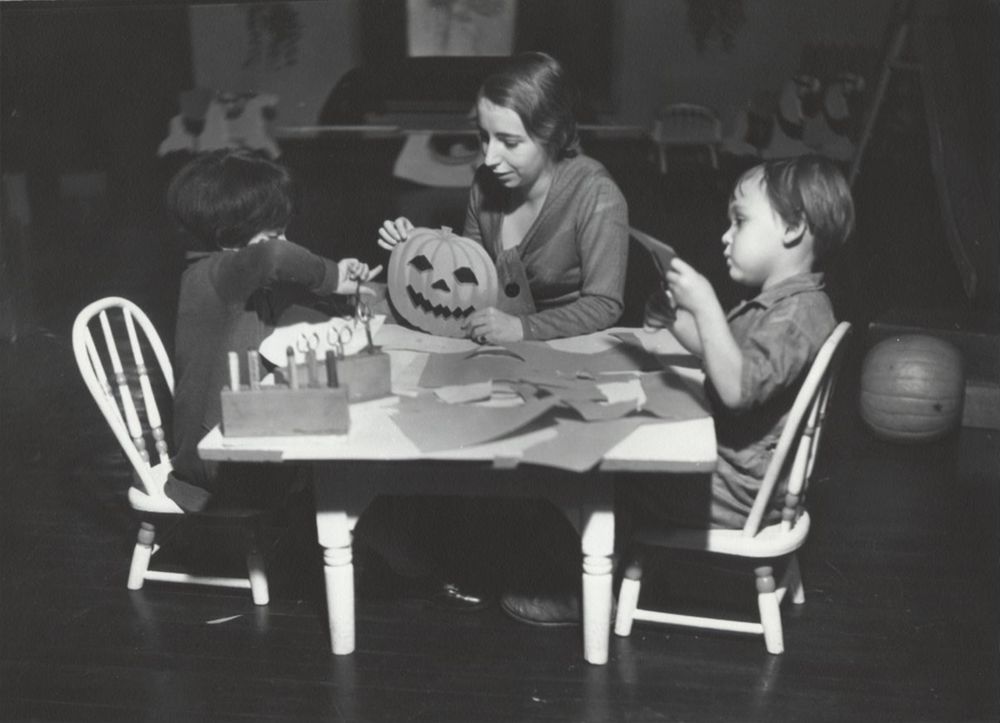 A woman and two children sit at a small table carving a jack-o-lantern together, surrounded by paper and craft supplies.