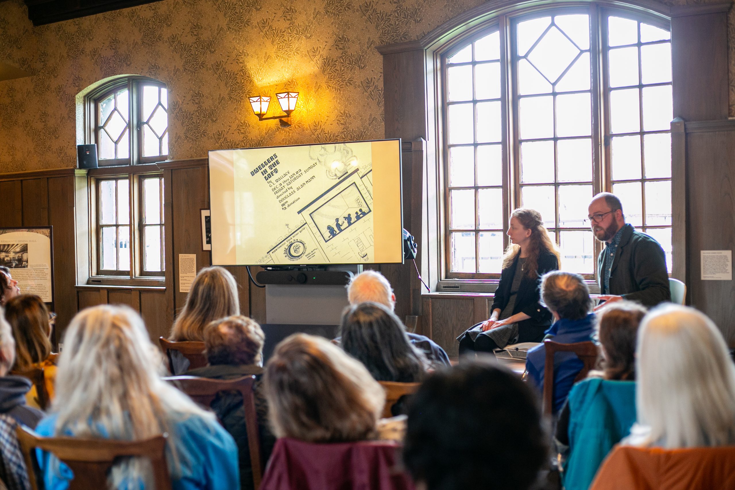 A group of people sit facing a screen and two presenters in a room with large windows and wooden walls, watching a presentation with diagrams and text on display.