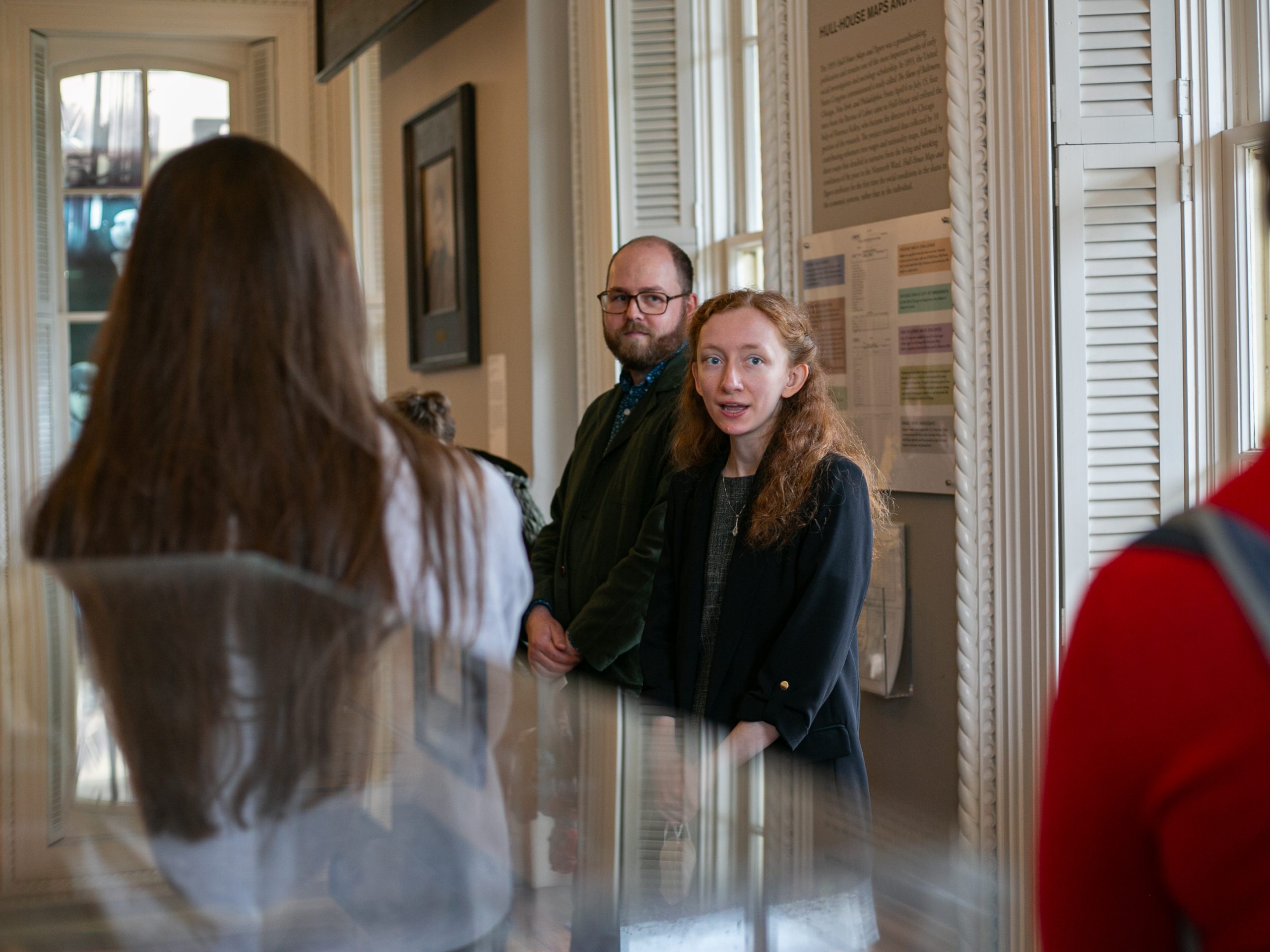 A woman and a man stand side by side, speaking to a small group of people inside a well-lit room with large windows and informational displays on the wall.