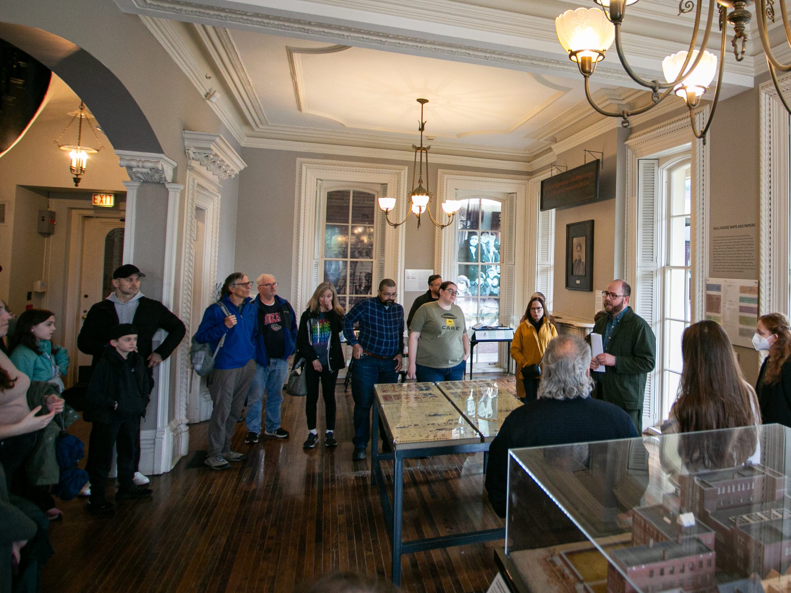 A group of people listens to a person speaking in a historic, well-lit museum room with wooden floors, chandeliers, and display cases. Some attendees stand, while others are seated or leaning against the wall.