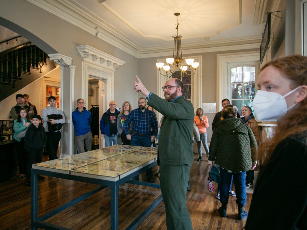 A tour guide gestures while speaking to a group of people inside a historic building. Some attendees listen attentively, and one person in the foreground wears a white face mask. The room features wooden floors and large windows.