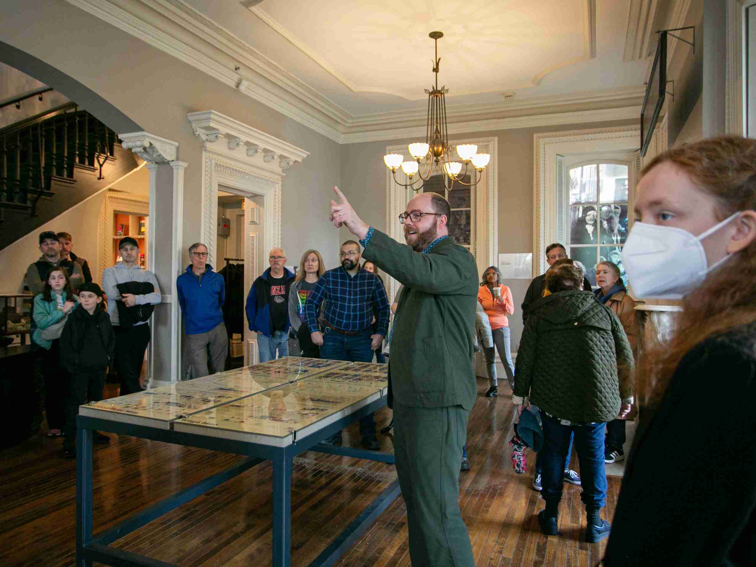A tour guide gestures while speaking to a group of people inside a historic building. Some attendees listen attentively, and one person in the foreground wears a white face mask. The room features wooden floors and large windows.