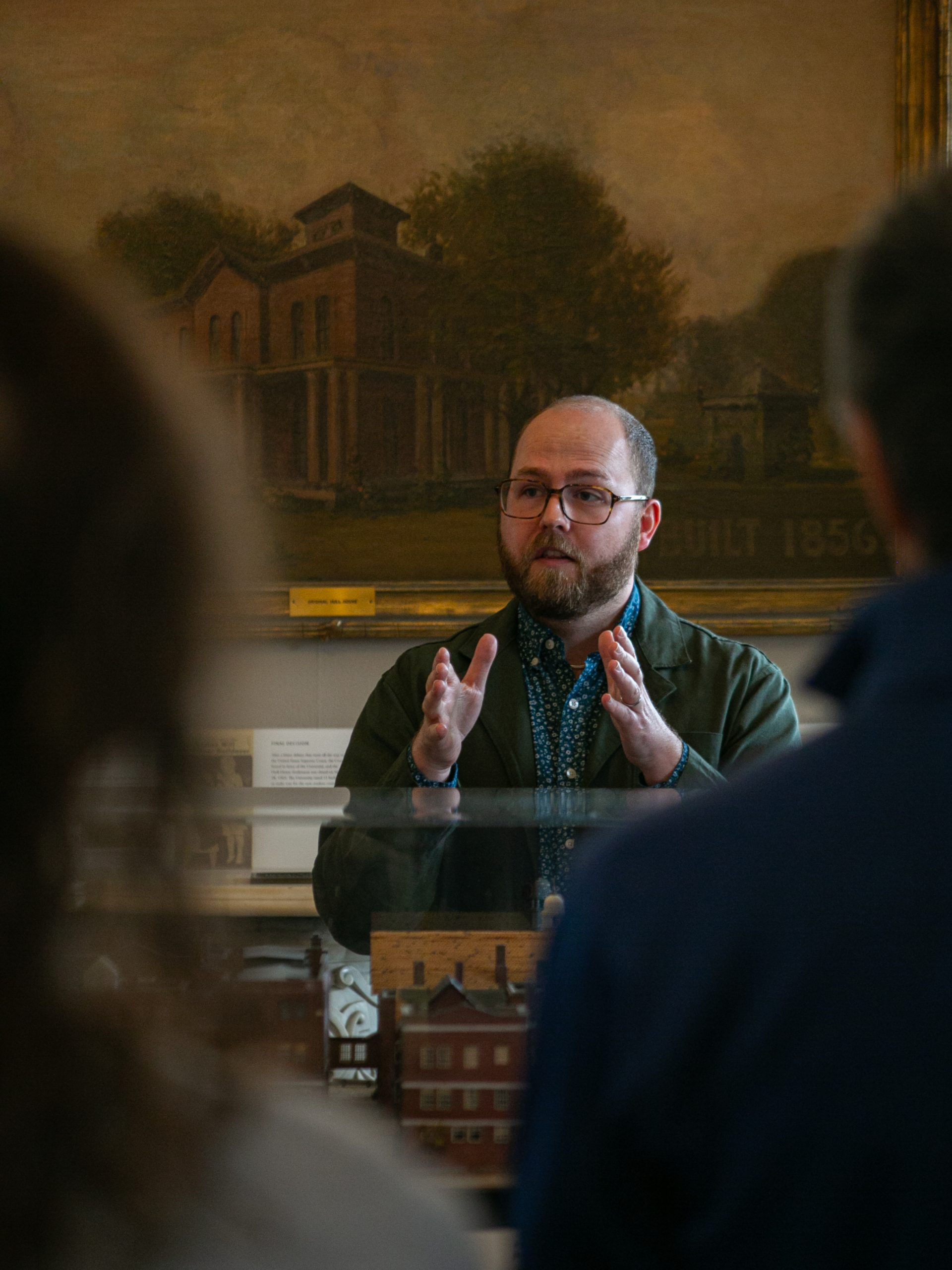 A man with glasses and a beard gestures while speaking to a small group in front of a display, with a painting of a house in the background.
