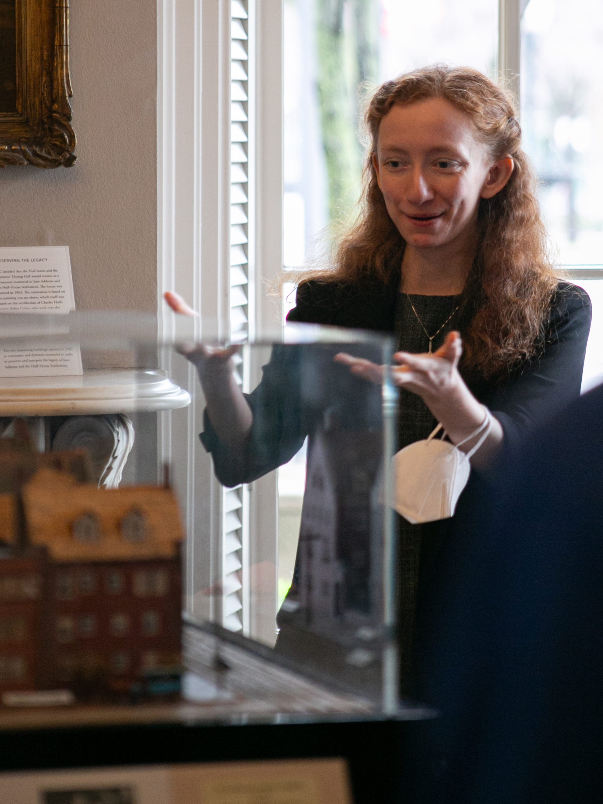 A woman with long red hair gestures enthusiastically while standing behind a glass display case containing a model building. She holds a white face mask in one hand, and natural light shines through a window behind her.