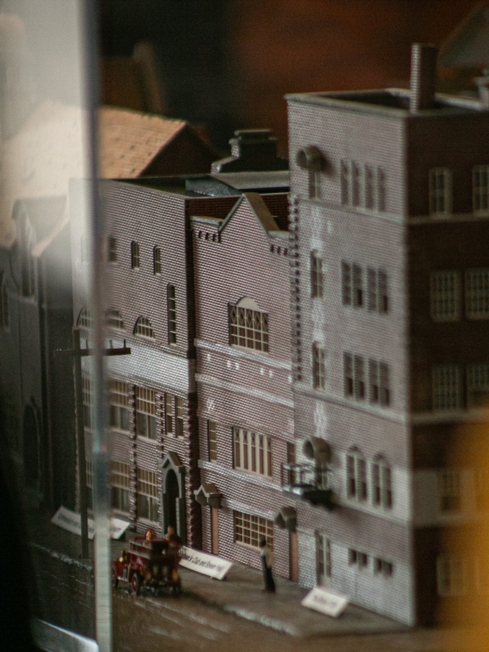 A close-up of a miniature model city block features detailed brick buildings and a small red fire truck on the street, viewed through a reflective glass surface.