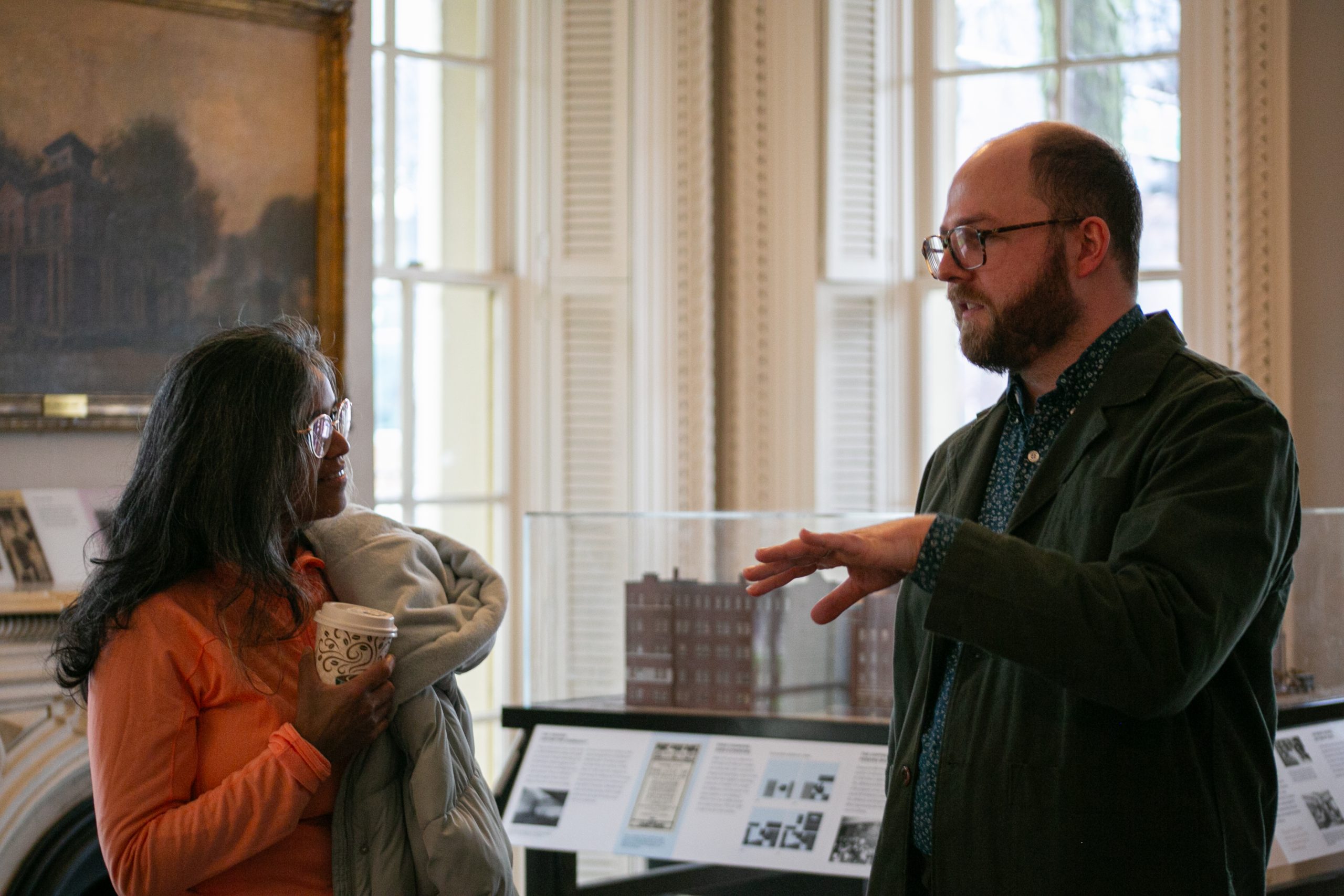 A woman holding a coffee cup listens to a man gesturing with his hand as they converse in a room with large windows, historical displays, and a framed painting on the wall.