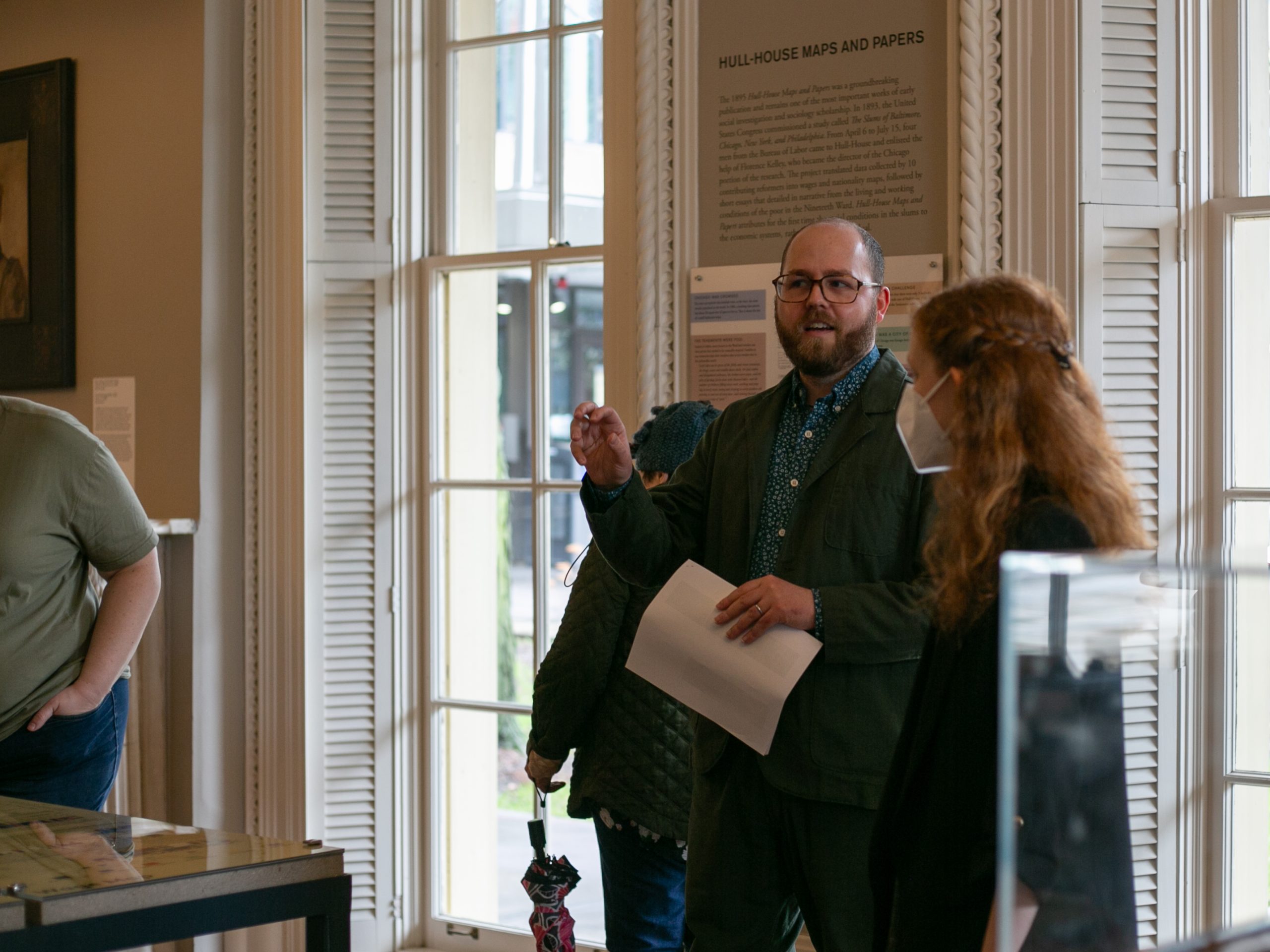 A person holding papers speaks to a small group inside a well-lit room with large windows and historical displays. One listener wears a face mask; others are partially visible.