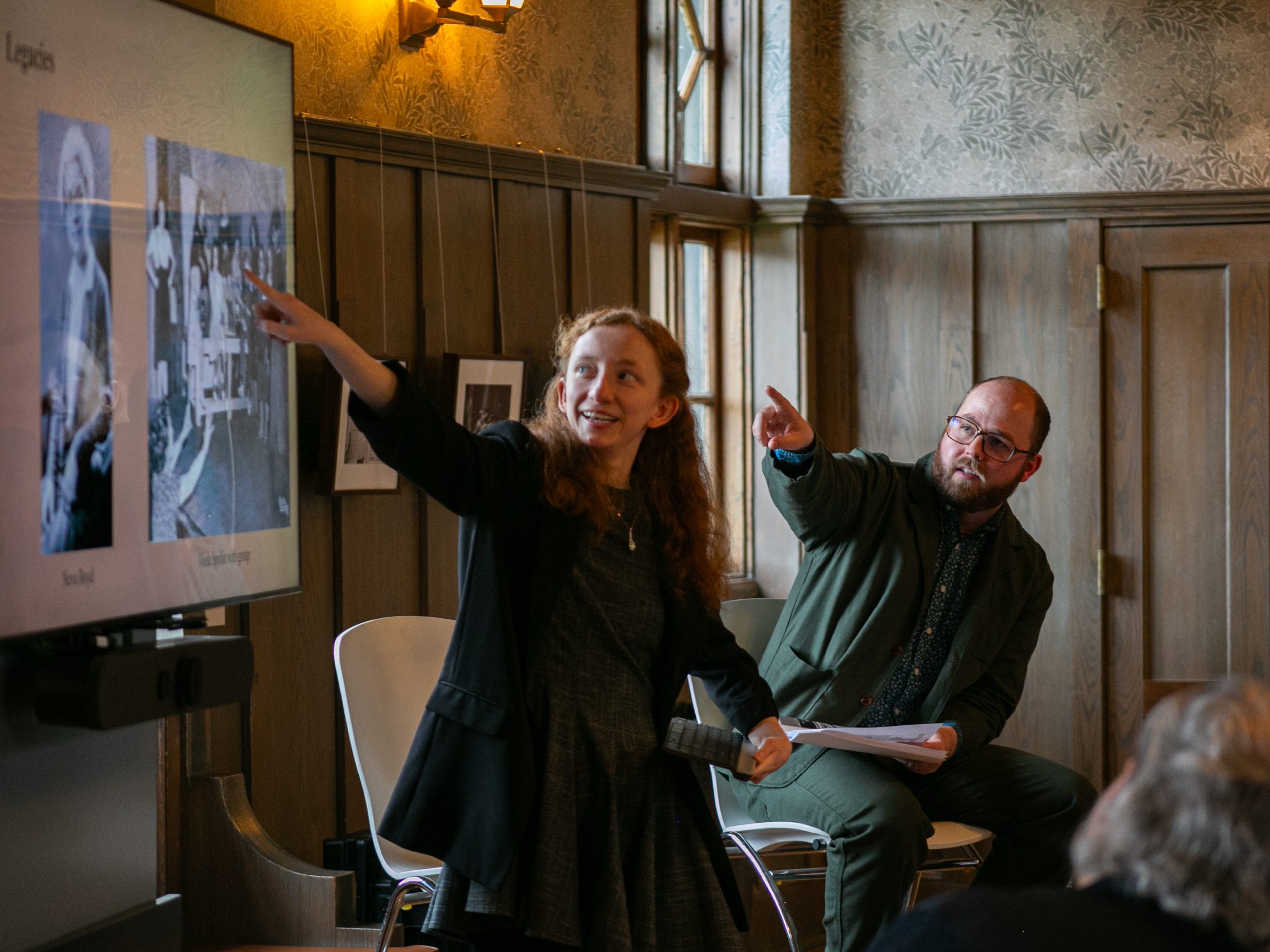 Two people sit side by side indoors, smiling and pointing toward a screen displaying historical black-and-white photos during a presentation. An audience is partially visible in the foreground.