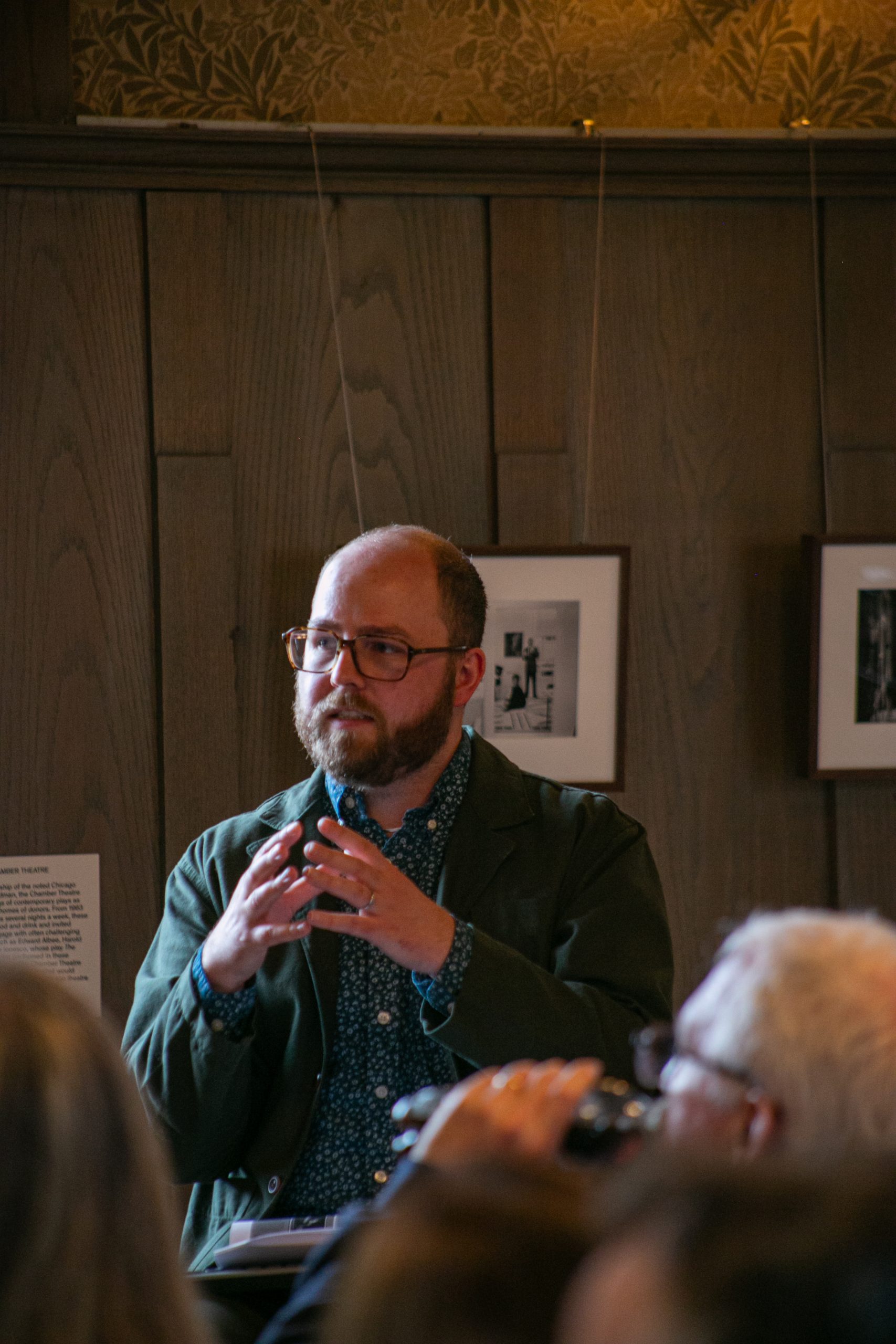 A bearded person with glasses and a patterned blue shirt speaks and gestures with their hands in a wood-paneled room, while others listen in the foreground. Framed photos hang on the wall behind them.