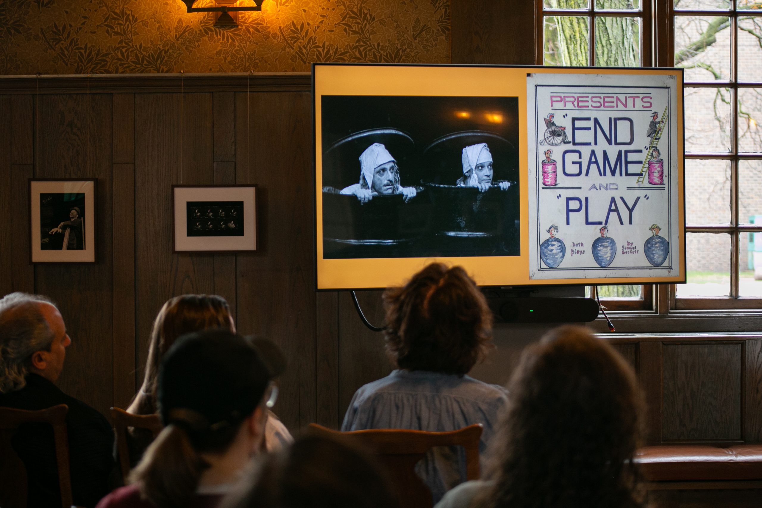 Several people sit facing a screen displaying a black-and-white scene of two actors in bins and a vintage poster reading End Game and Play. The setting appears to be a wood-paneled room with framed photos on the wall.