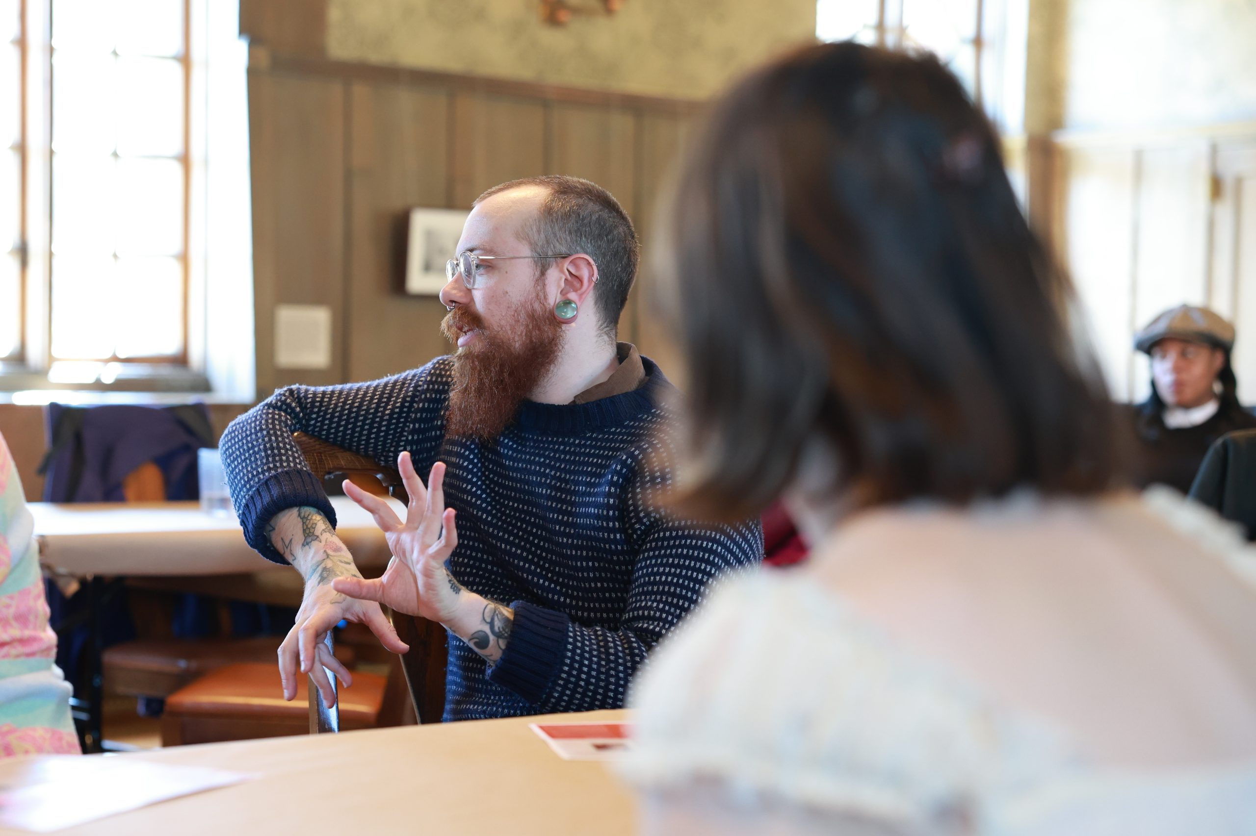 A bearded person with tattoos and glasses gestures while speaking at a table in a well-lit room, with another person in the foreground and others in the background.