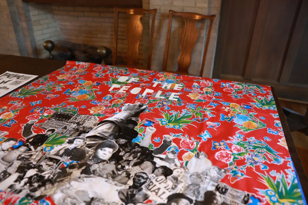 A colorful collage with “OF THE PEOPLE” text is spread on a table, featuring black-and-white photos of people and flowers on a red floral background. Two wooden chairs and a brick fireplace are in the background.