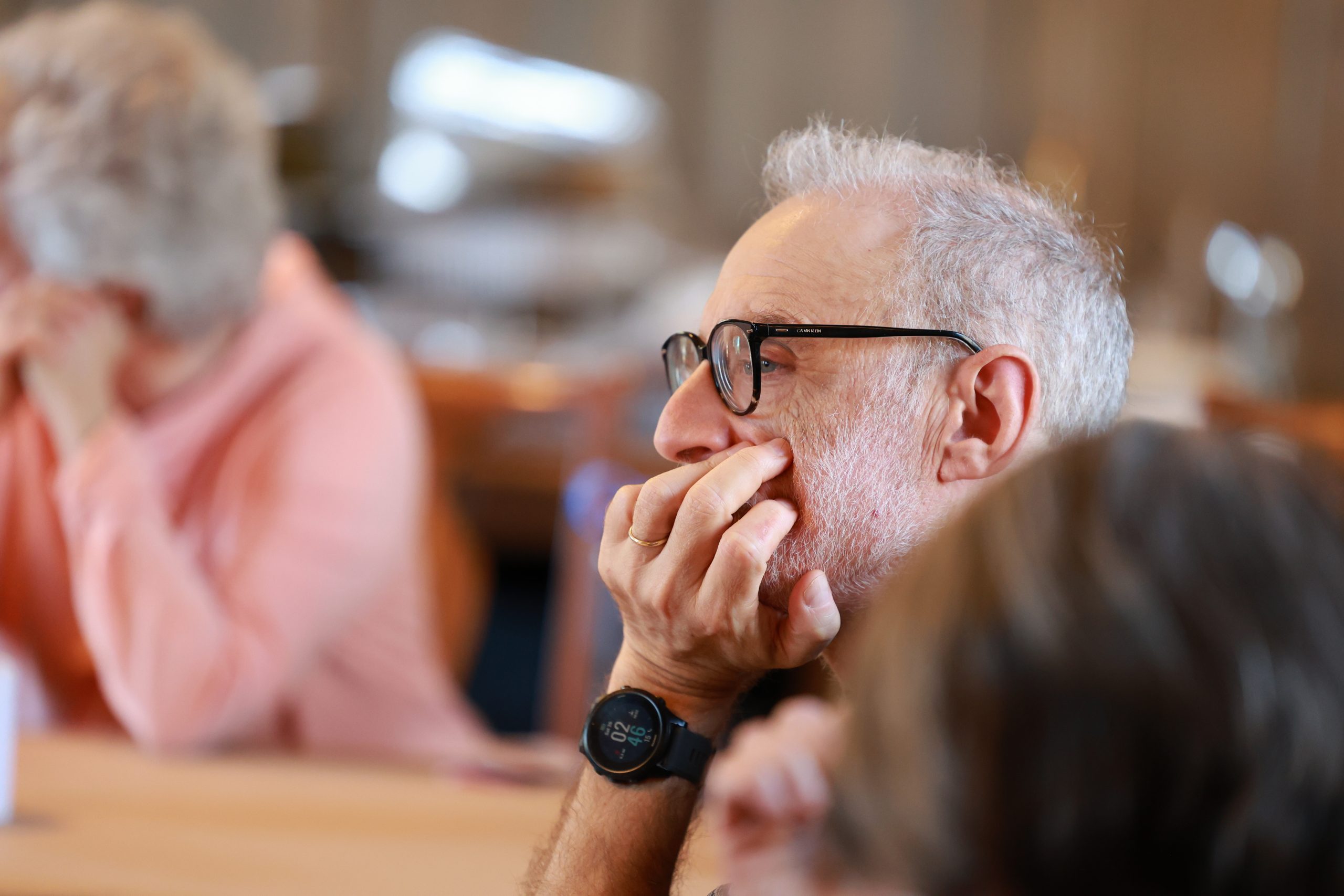 A man with gray hair and glasses, wearing a black wristwatch, rests his chin on his hand while listening attentively in a blurred indoor setting with other people in the background.
