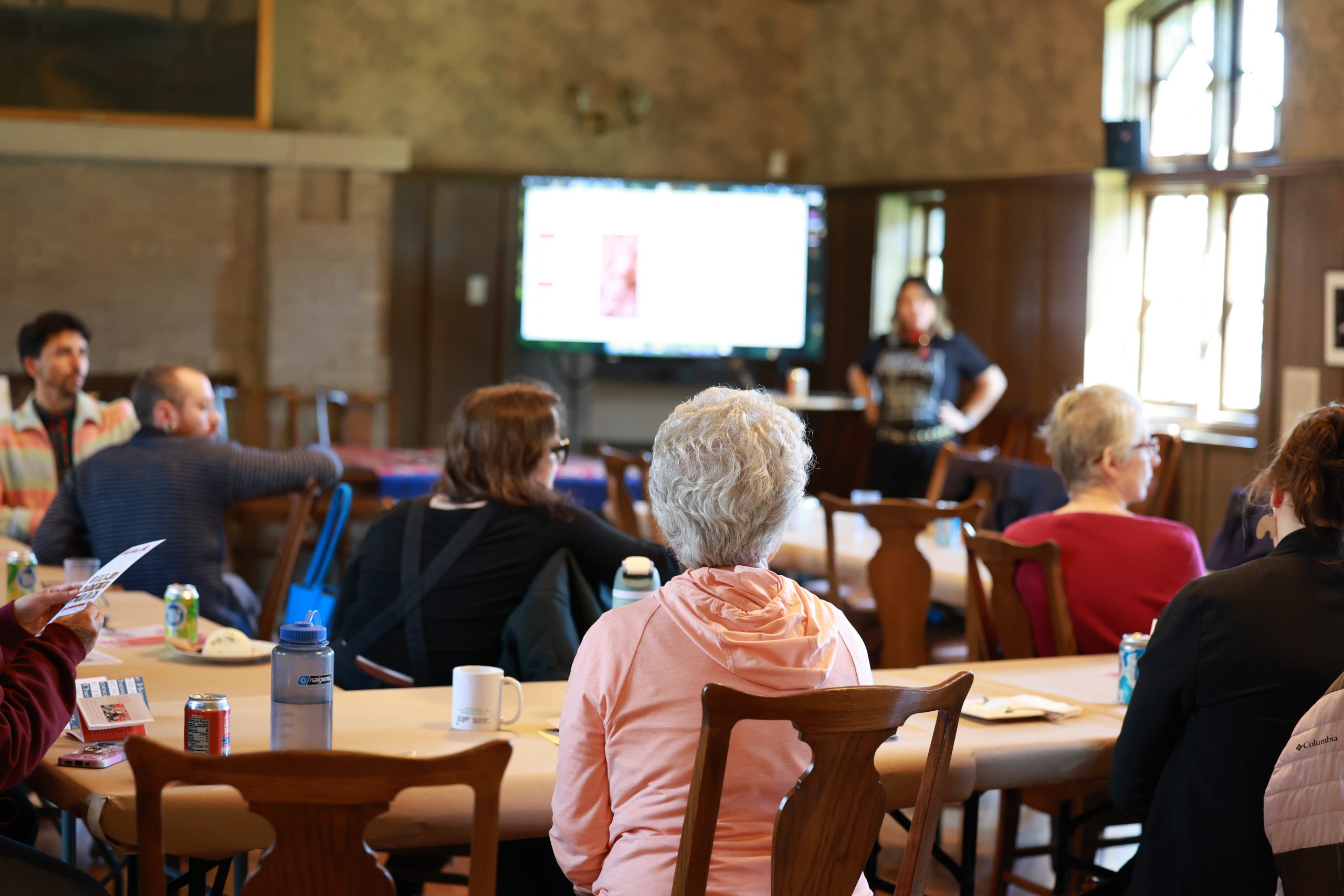 People sit at tables in a warmly lit room, facing a woman presenting near a screen. Drinks and papers are on the tables, and sunlight streams through the windows on the right side of the room.