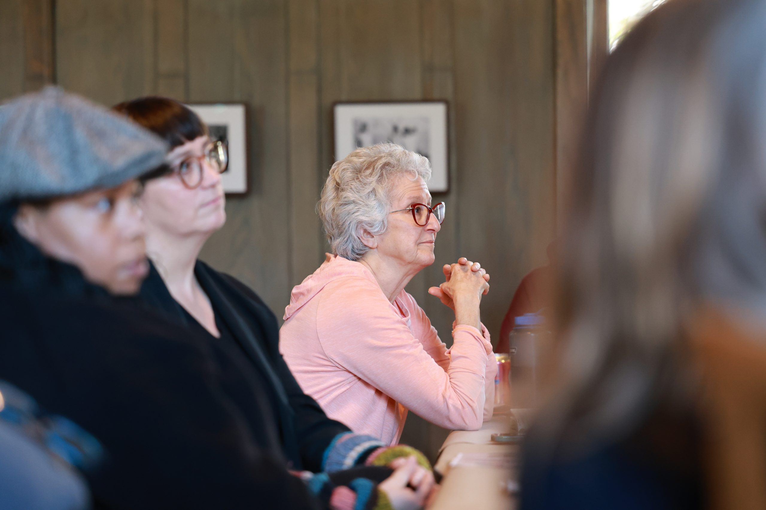 A group of people sits attentively indoors, focusing on something out of view. A woman with gray hair and glasses, wearing a pink sweater, is in focus and appears to be listening closely.
