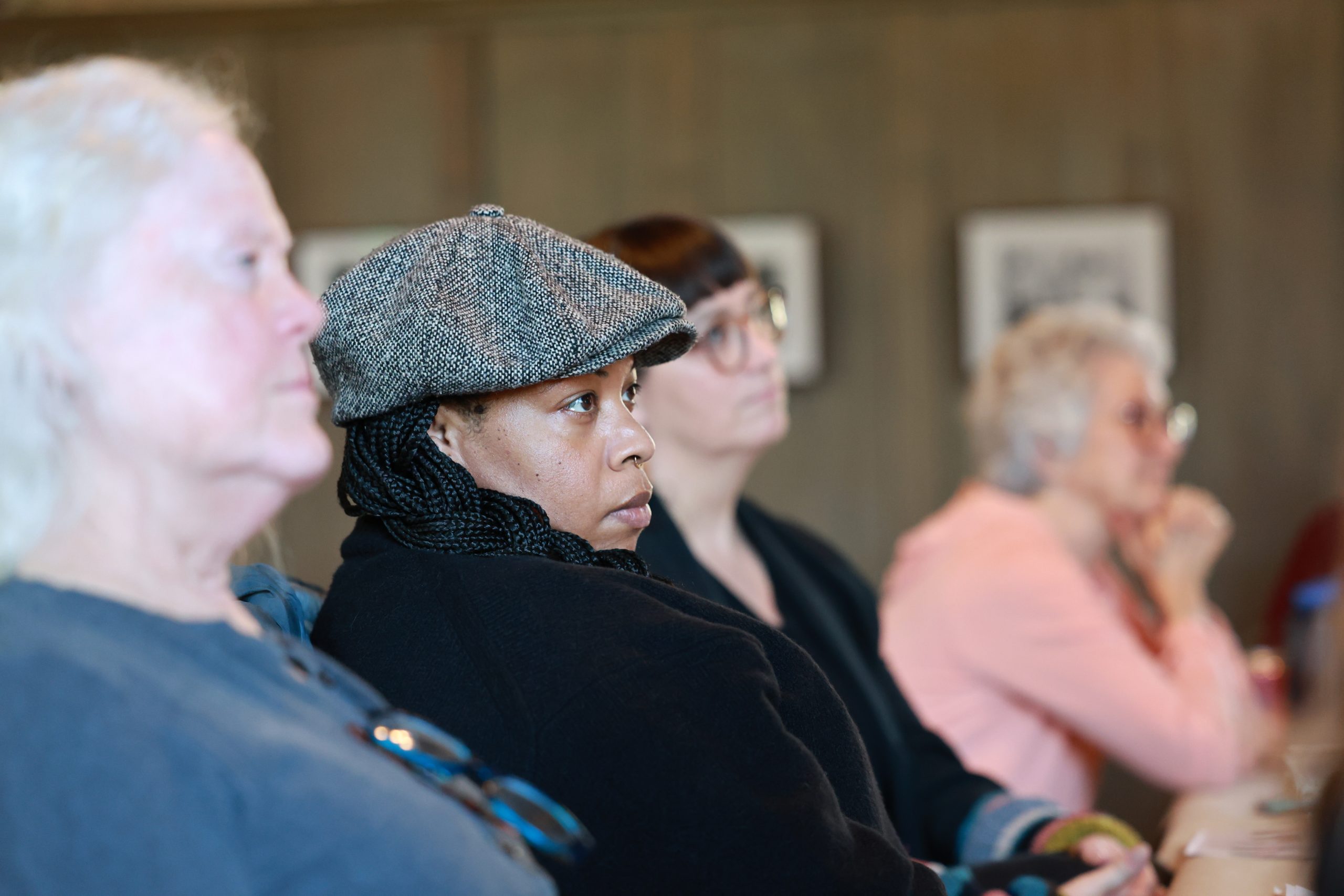 A group of people sit in a row, attentively listening. The focus is on a person in the center wearing a gray cap and a black jacket, with others beside them slightly out of focus.