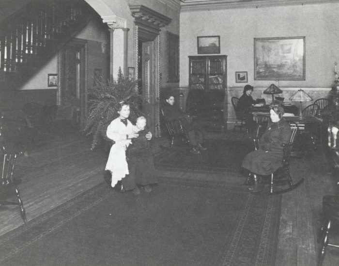 Black and white photo of a vintage room with five people sitting or standing, wooden furniture, lamps, a staircase on the left, and framed art on the walls. Two children stand together in the foreground.