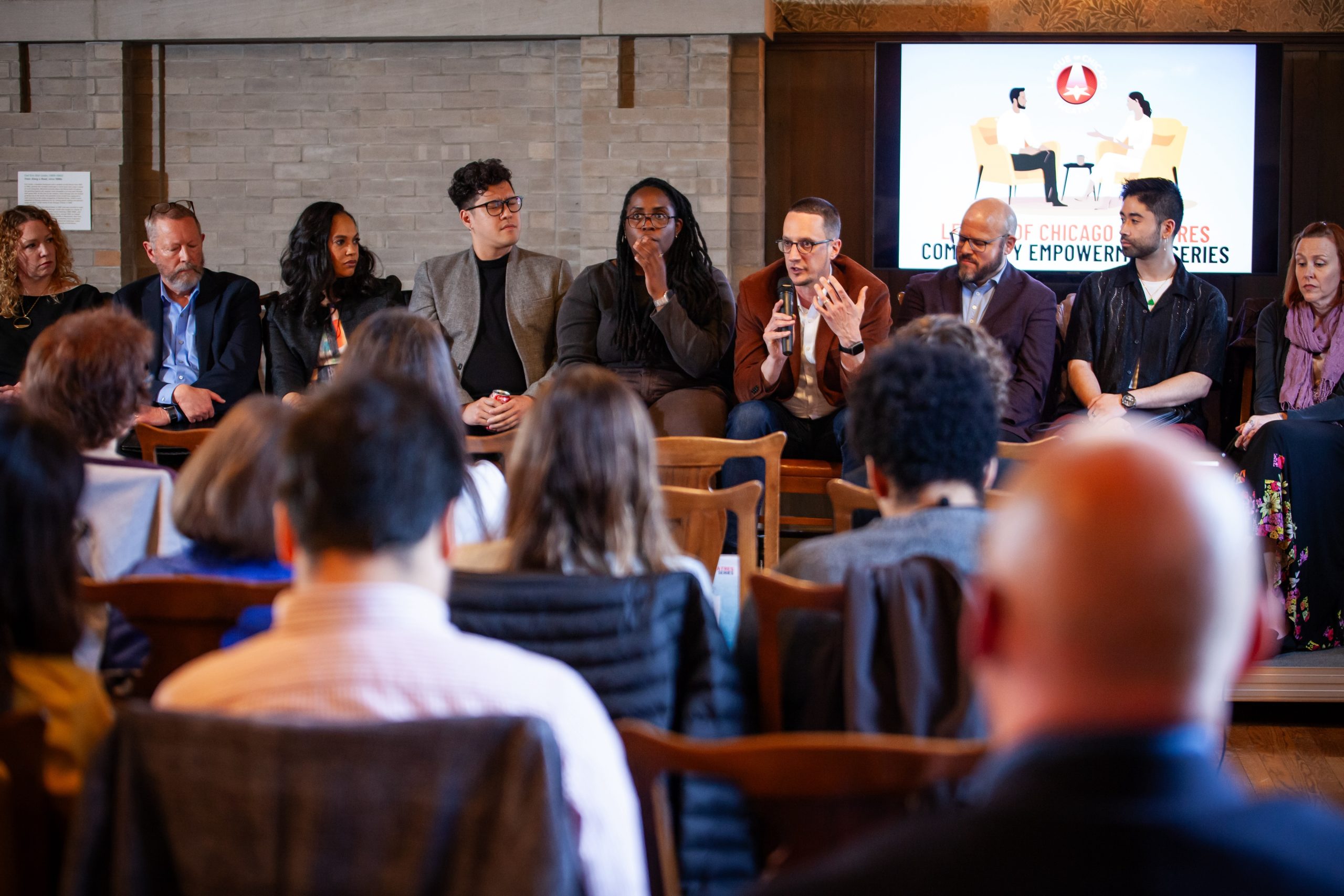 A diverse panel of speakers sits in front of an audience during a discussion event. One panelist speaks into a microphone while others listen. A presentation screen is visible in the background.