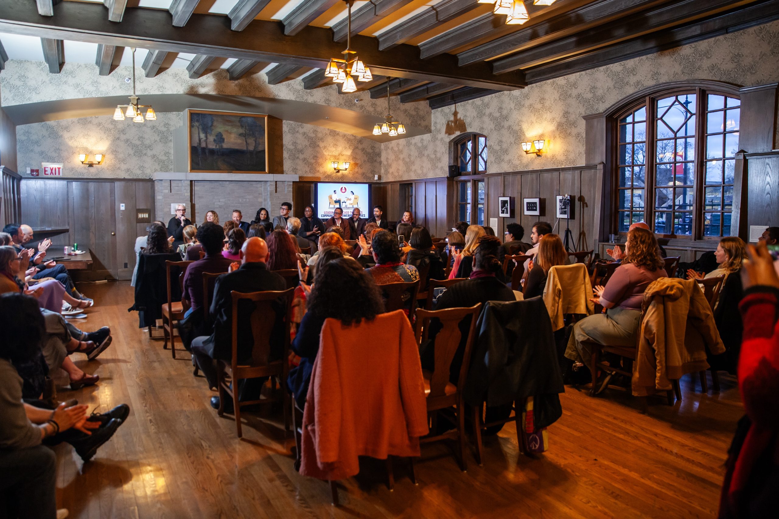 A group of people sit in a rustic, wood-paneled room with large windows, attending a panel discussion or presentation at the front where several speakers are seated beside a screen.
