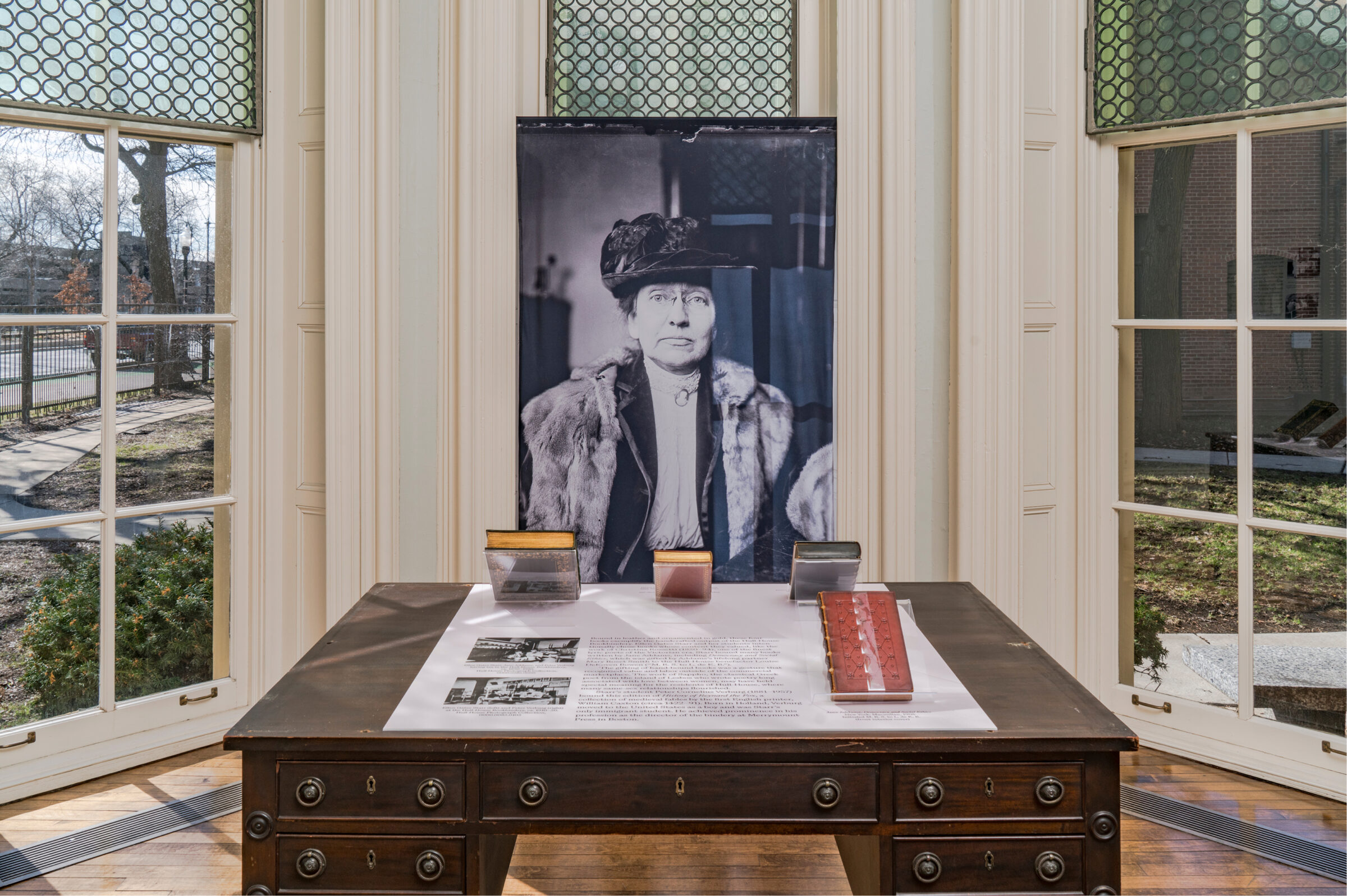 A historical exhibit on a wooden desk features a large black-and-white portrait of a woman in a fur coat and hat, surrounded by documents and display cases, set in front of tall windows.