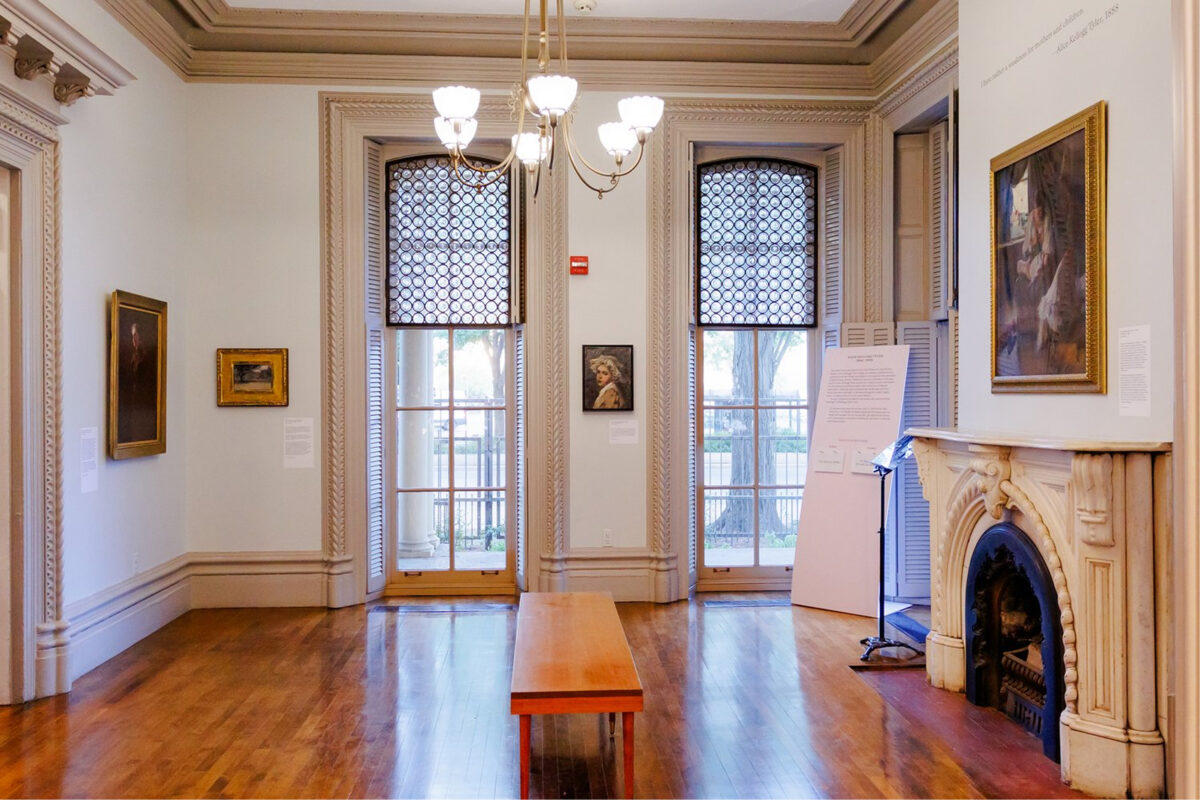 A bright, elegant art gallery room with wooden floors, a bench in the center, a chandelier, ornate fireplace, large windows with patterned shades, and several framed paintings on the walls.