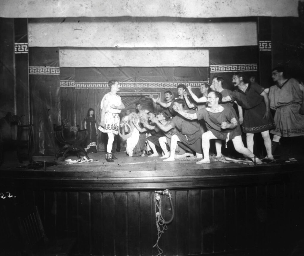 A group of people in tunics and sandals perform an energetic scene on stage; several reach toward a person standing apart, under theatrical lighting, with a backdrop featuring Greek-style patterns.