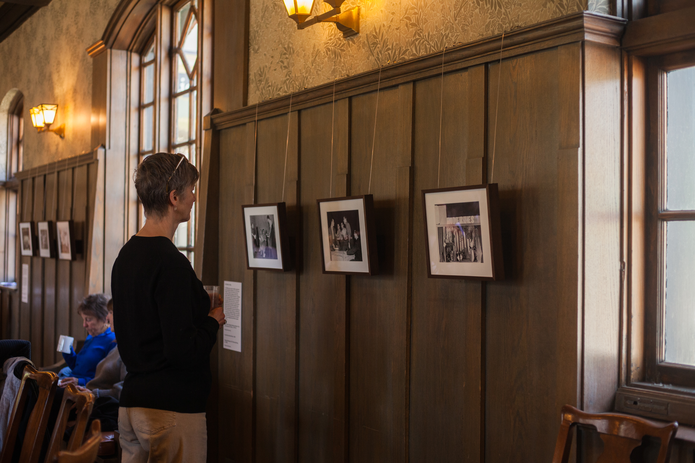 A person stands in a wood-paneled room, looking at framed photographs displayed on the wall. Other people are seated nearby, and natural light comes through large windows.
