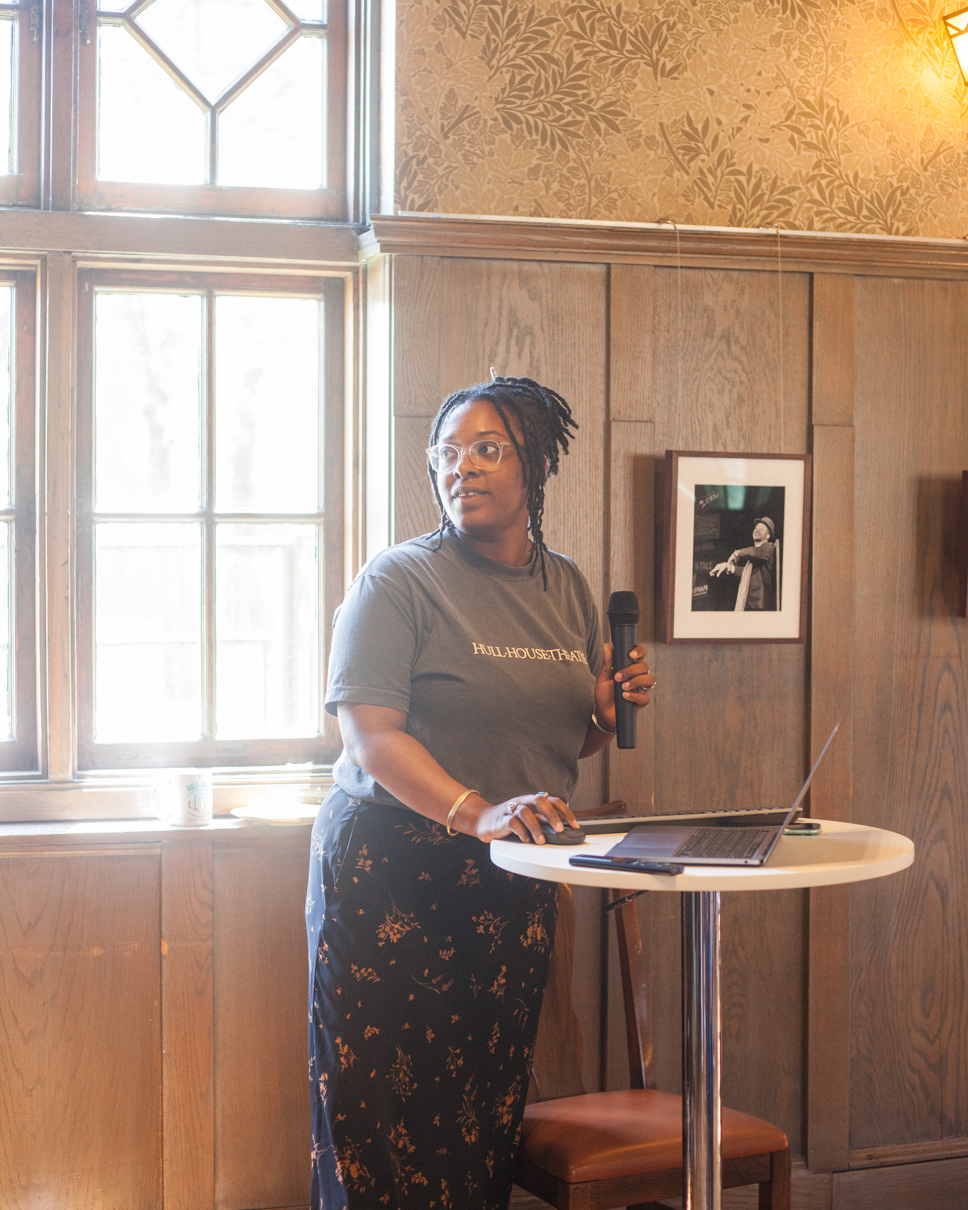 A person stands at a small round table with a laptop, holding a microphone and looking to the side. Sunlight streams through large windows behind them. A framed photo and patterned wallpaper decorate the wooden wall.
