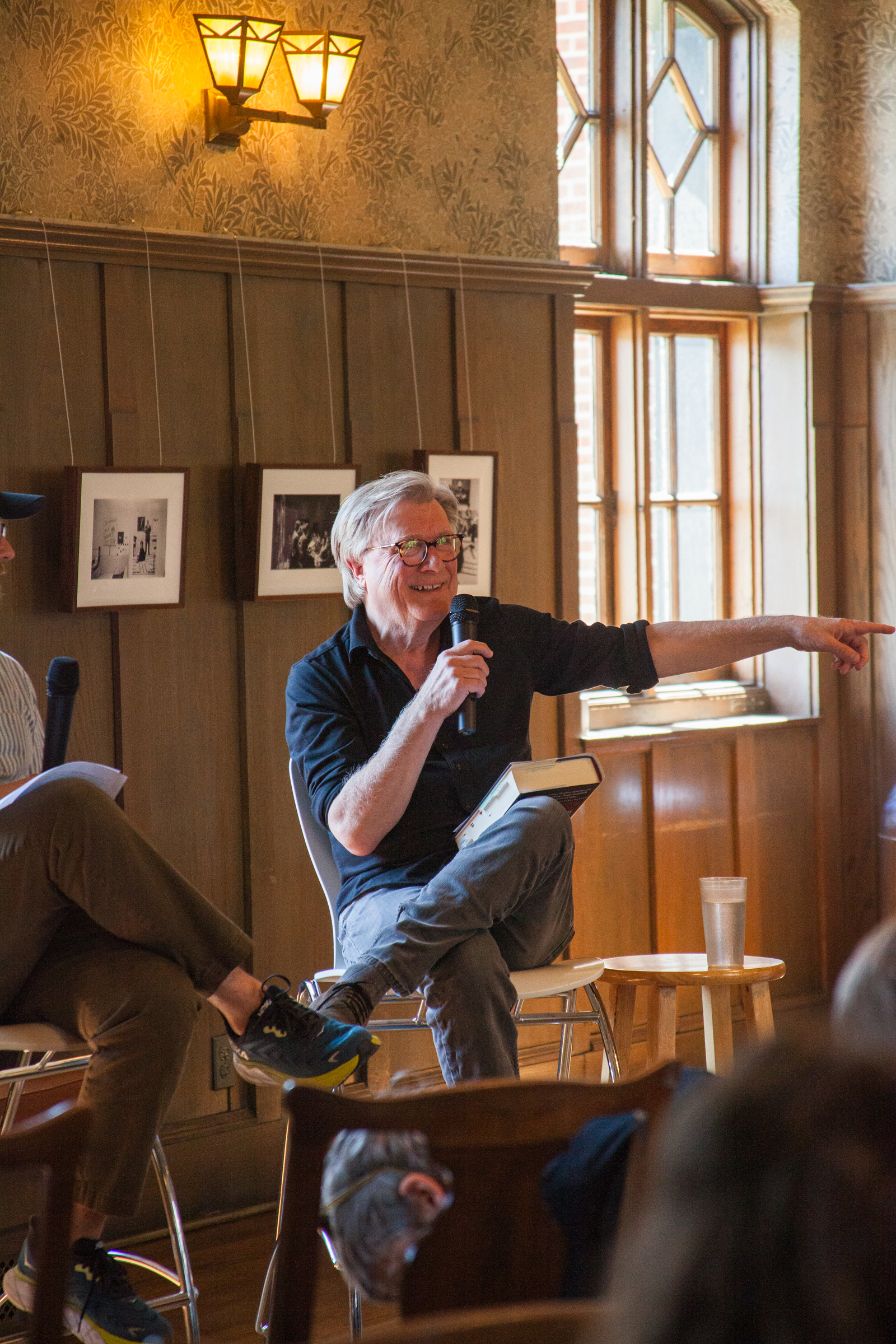 A smiling older man with glasses sits on a stool, holding a microphone and pointing, while talking to an audience in a warmly lit room with wooden walls, framed photos, and tall windows.