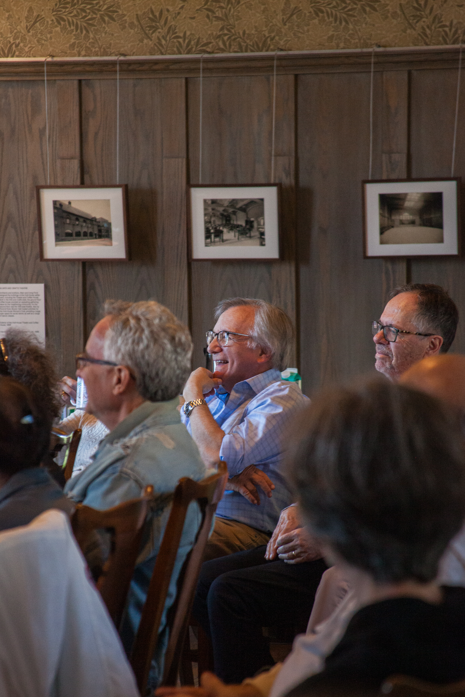 A group of people sit indoors, attentively listening and smiling during an event. Framed black-and-white photos hang on the wooden-paneled wall behind them. The atmosphere appears warm and engaging.