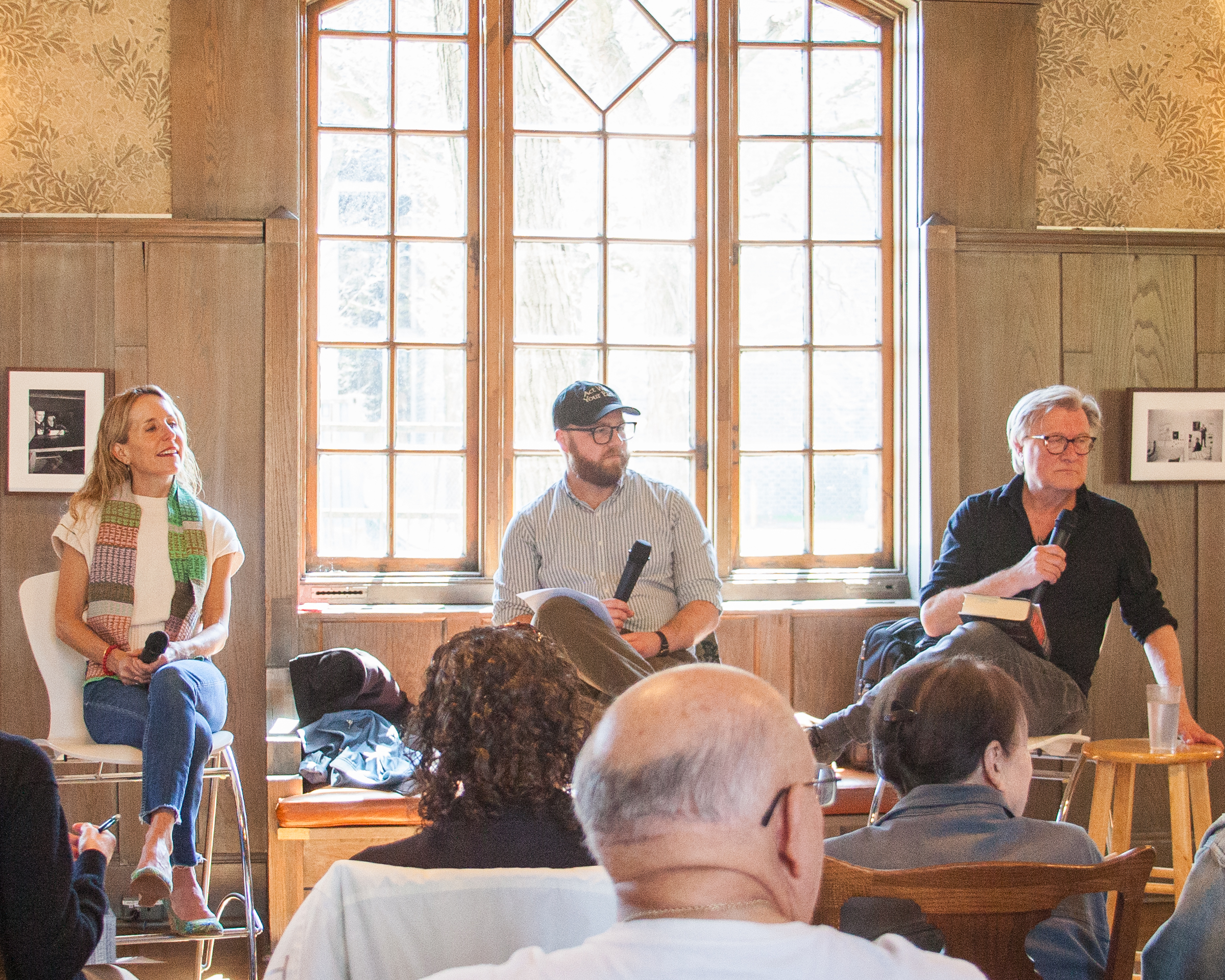 Three people sit on chairs at the front of a room, holding microphones, facing an audience. Sunlight streams through tall windows behind them. The audience listens attentively. The setting looks informal and inviting.