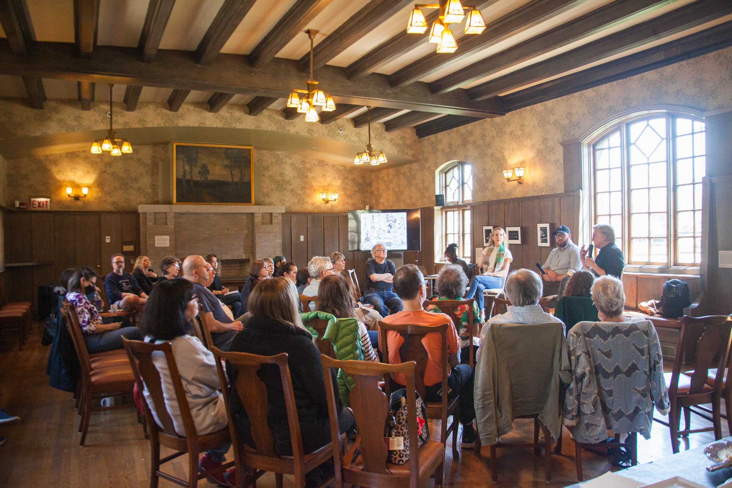 A group of people sit in a well-lit, wood-paneled room with large windows, listening to a speaker at the front. The audience appears engaged and the atmosphere is warm and inviting.