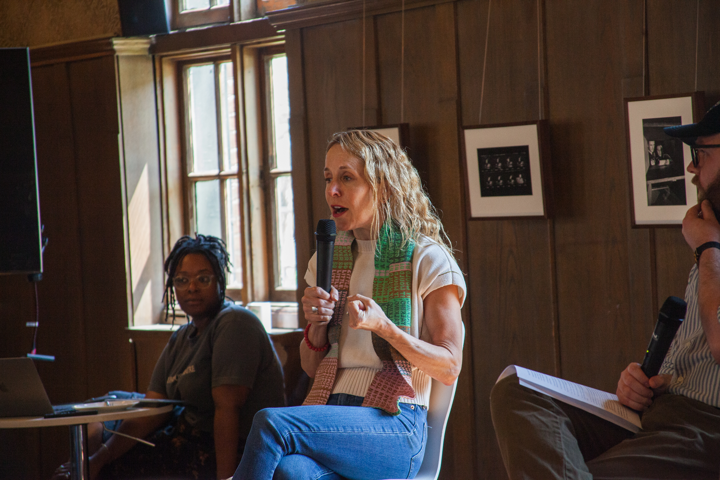 A woman holding a microphone speaks while seated on a panel indoors, next to two other people. She gestures with her hand. Framed photos and a window are visible on the wood-paneled wall behind them.