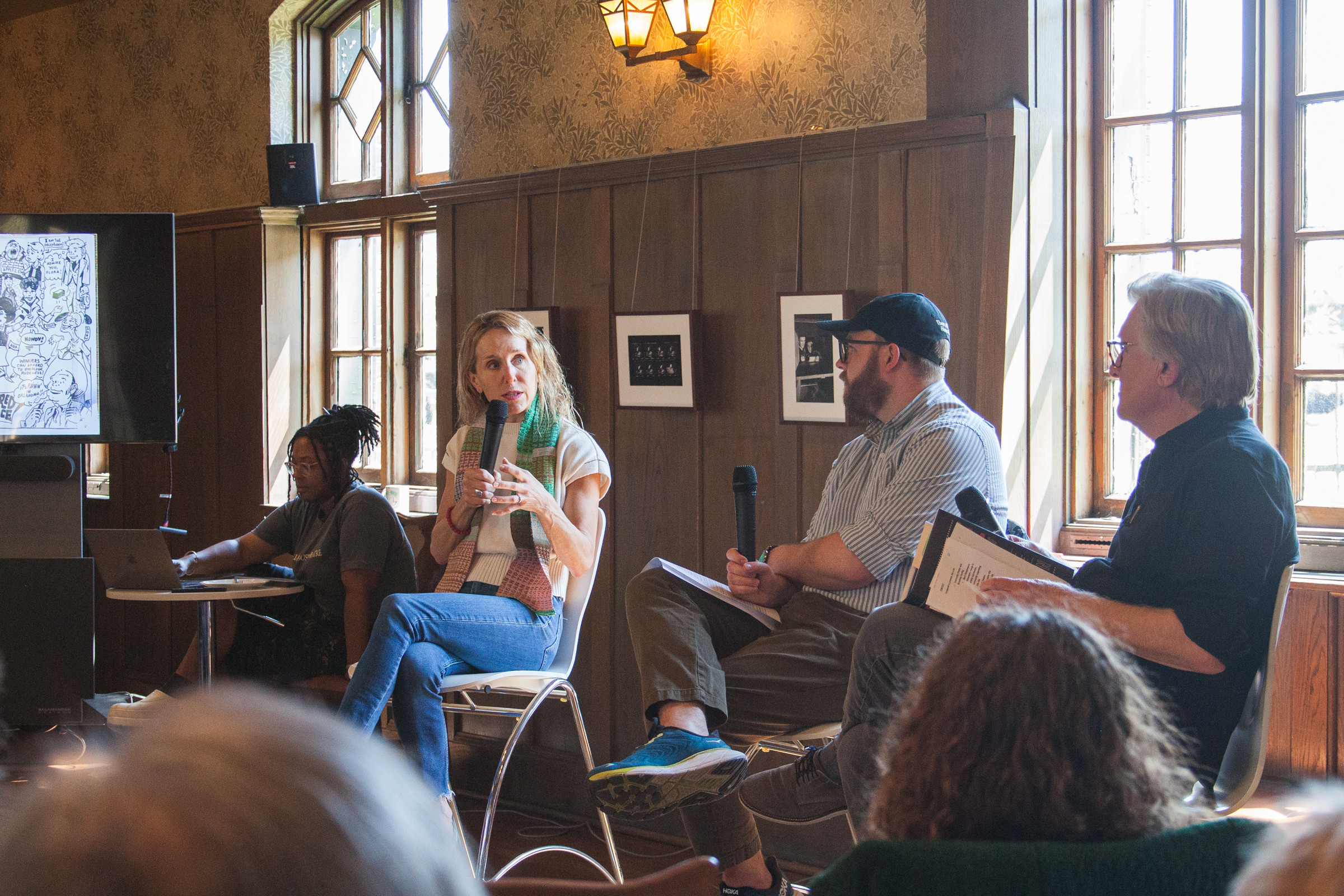 Four people sit on a panel in a wood-paneled room with large windows. One woman speaks while holding a microphone, and two men beside her also hold microphones. Another woman sits at a table with a laptop. Audience members are visible in front.