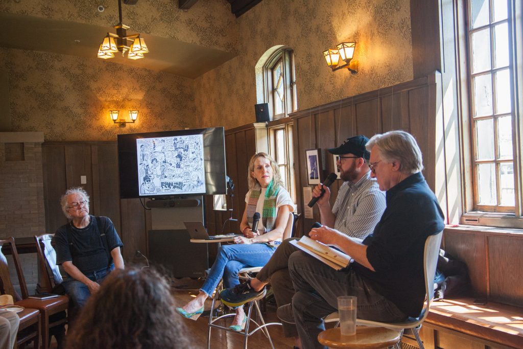 Four people sit at the front of a room for a panel discussion. One man speaks into a microphone, while a woman and two other men listen. A screen behind them displays black-and-white illustrations. Audience members watch.