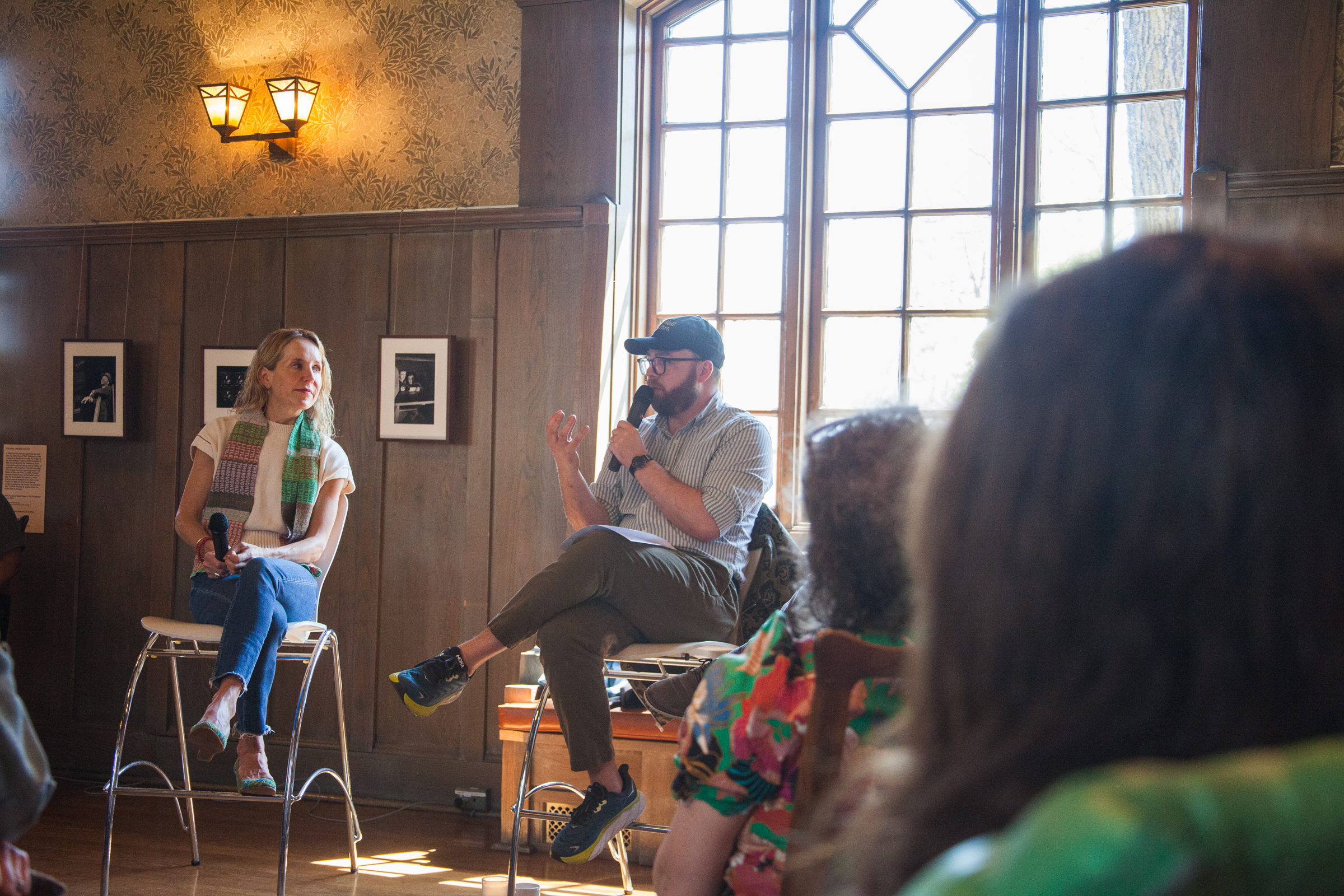 A man and woman sit on stools speaking to an audience in a sunlit room with wood-paneled walls and large windows. The woman listens while the man gestures with his hand, holding a microphone.