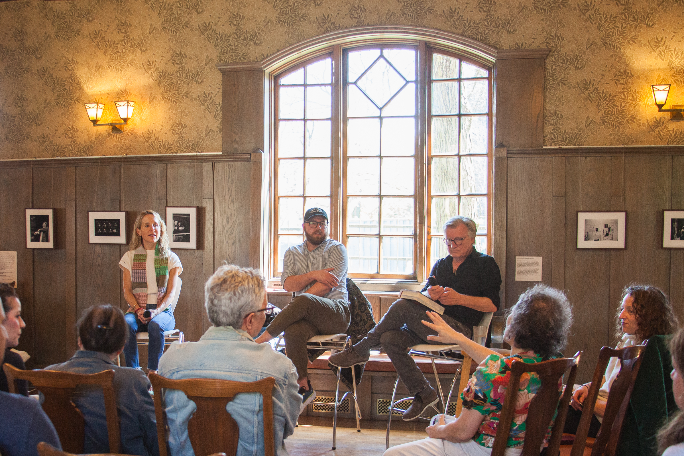 A group of people sit in a wood-paneled room with a large window. Three people are seated at the front, engaging with the audience, who are seated in chairs facing them. Black-and-white photos hang on the walls.