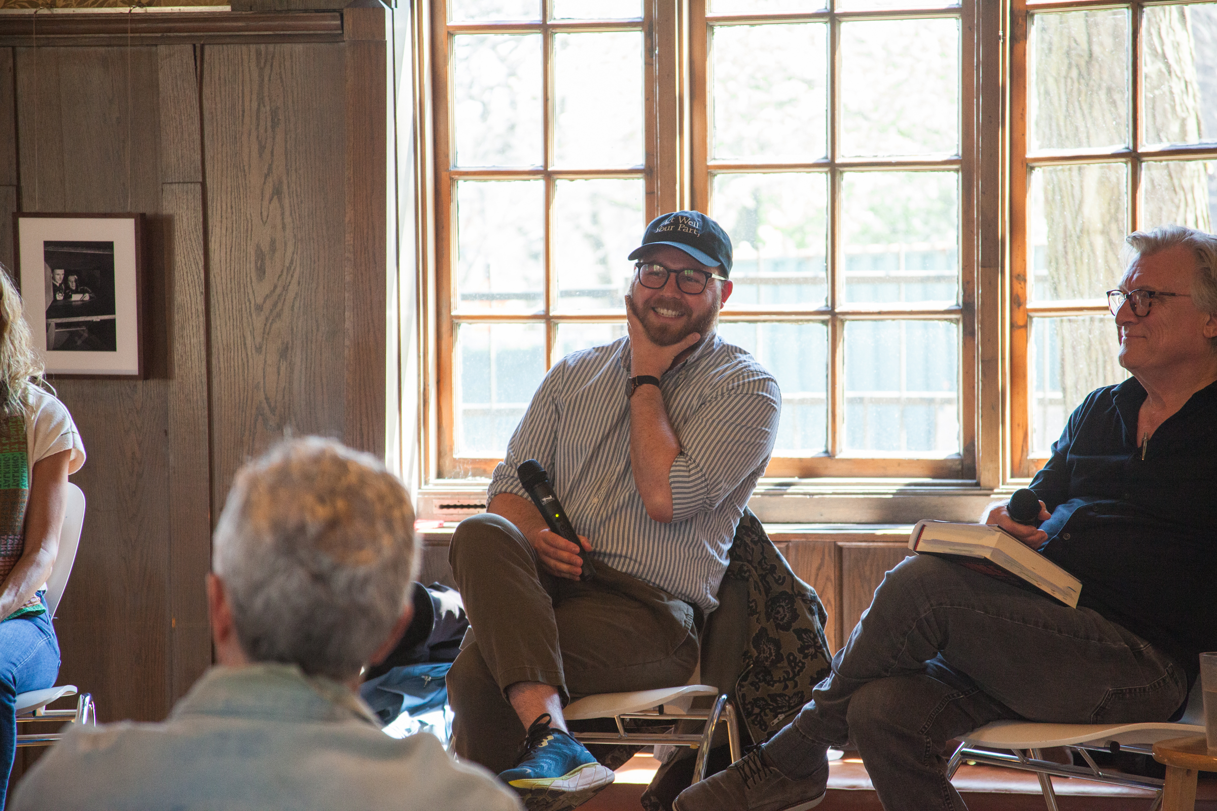 A man in a baseball cap and glasses smiles while holding a microphone, seated next to another man with a book. They are in a bright room with wood-paneled walls and large windows. People are seated and listening.