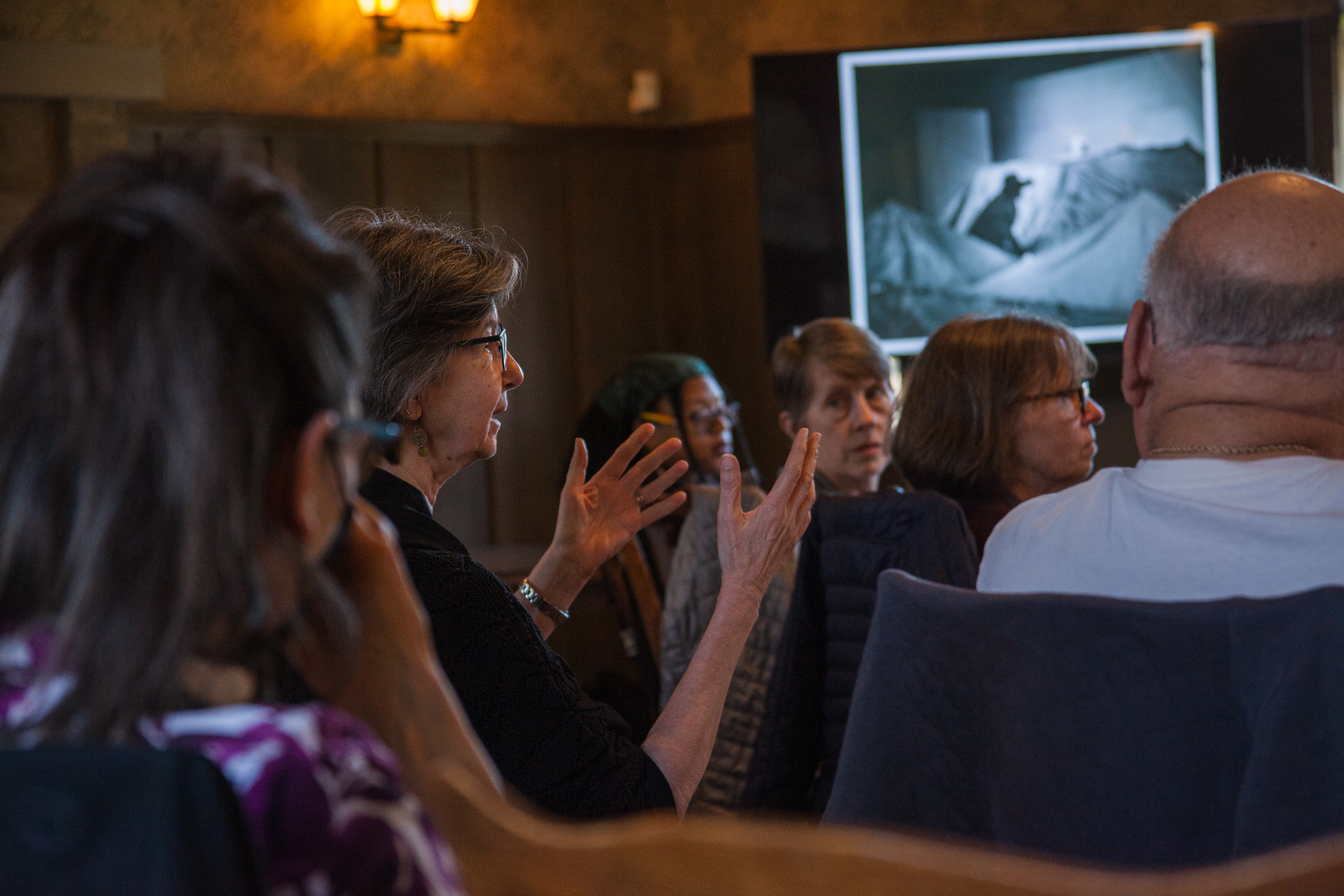 A woman gestures while speaking in a dimly lit room with several people seated around her. A black-and-white photo is displayed on a screen in the background.