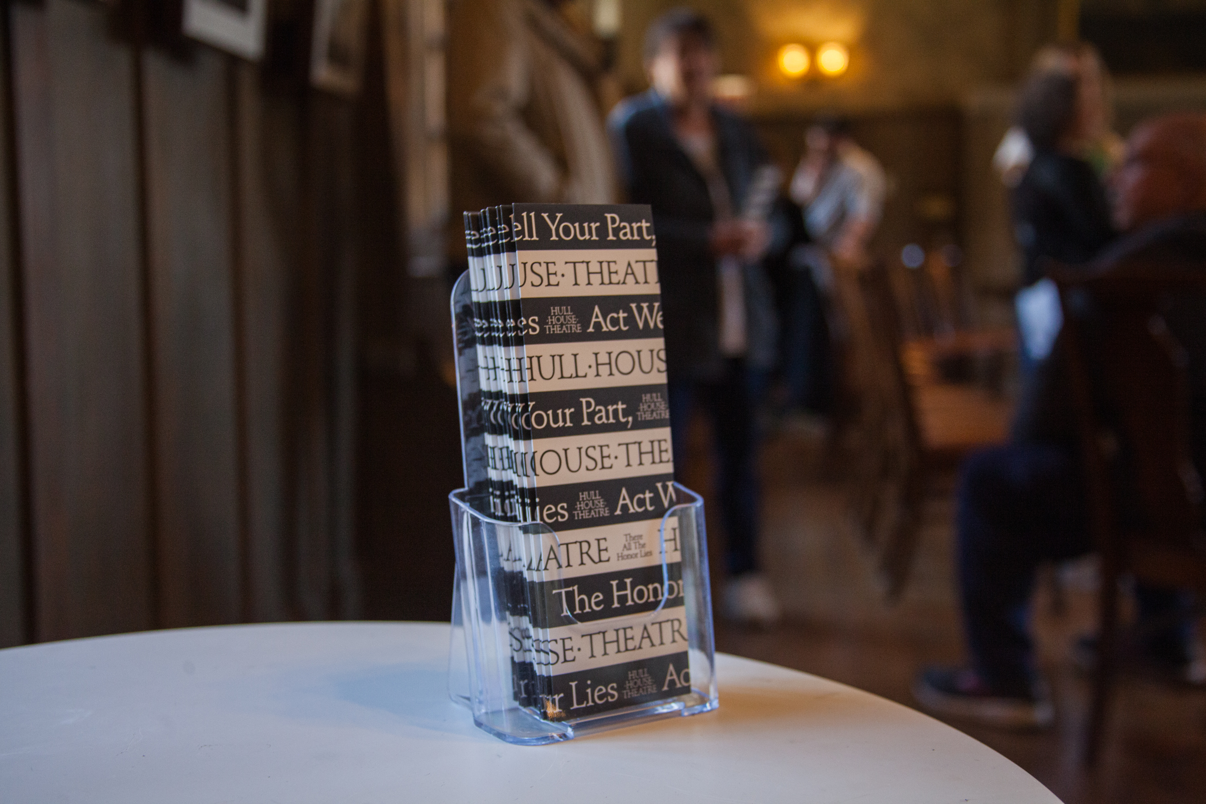 A clear plastic holder on a white table displays brochures labeled Hull-House Theatre. Several people stand blurred in the background inside a warmly lit room with wooden walls.