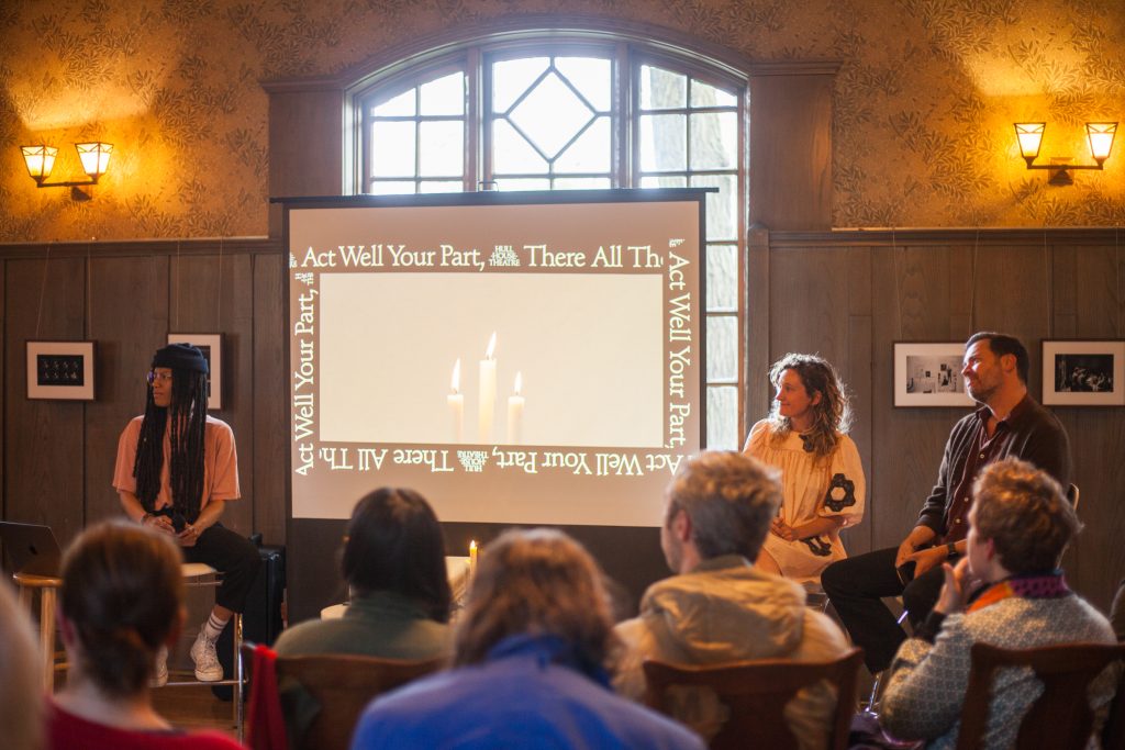 A group of people listens to three speakers seated in front of a projected screen in a warmly lit room. The screen displays candles and the phrase Act Well Your Part; There All The Honor Lies.