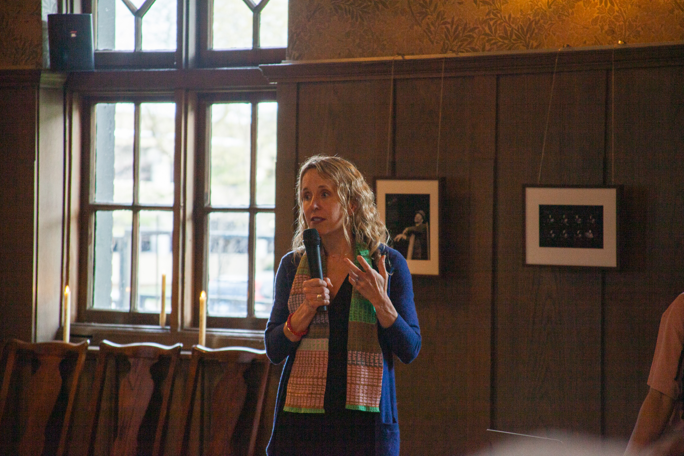 A woman with curly blonde hair speaks into a microphone in a wood-paneled room with large windows and framed photos on the wall. She wears a colorful scarf and gestures with one hand while talking.