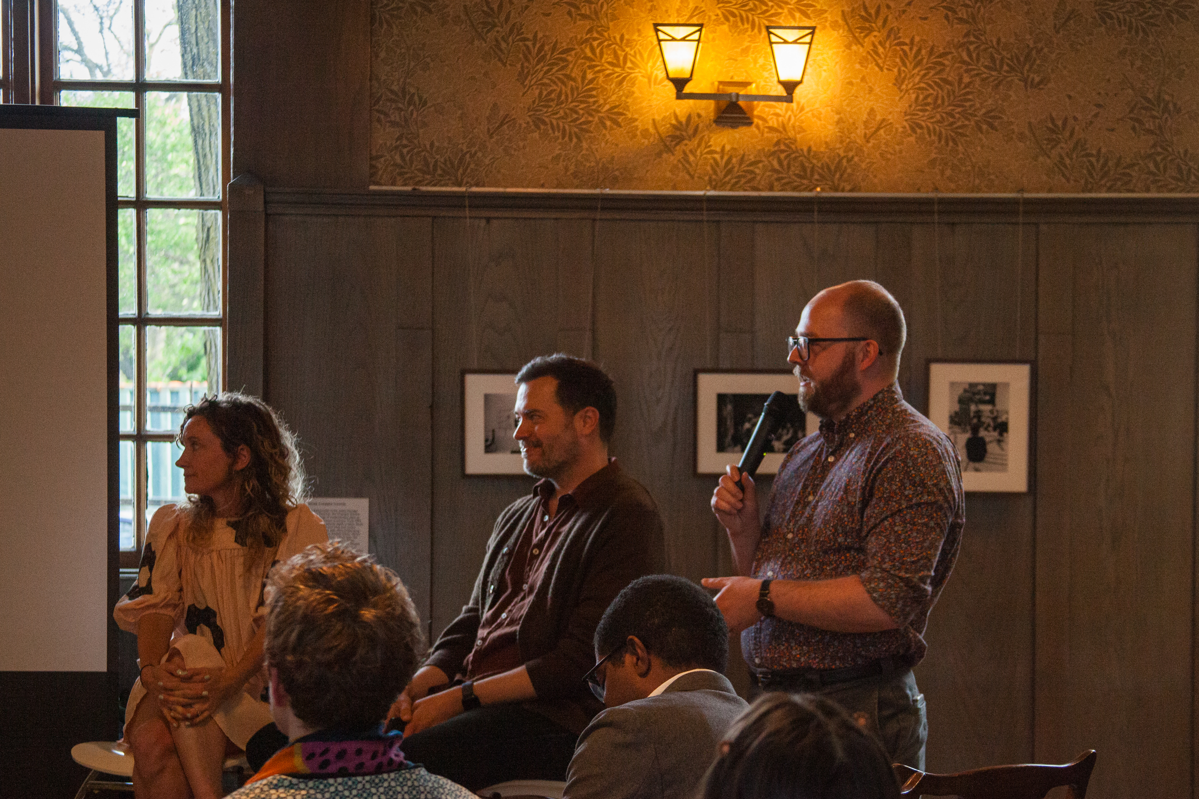 A man holding a microphone speaks while standing beside a seated man and woman during an indoor panel discussion; audience members are visible in the foreground. Warm lighting and framed photos decorate the room.