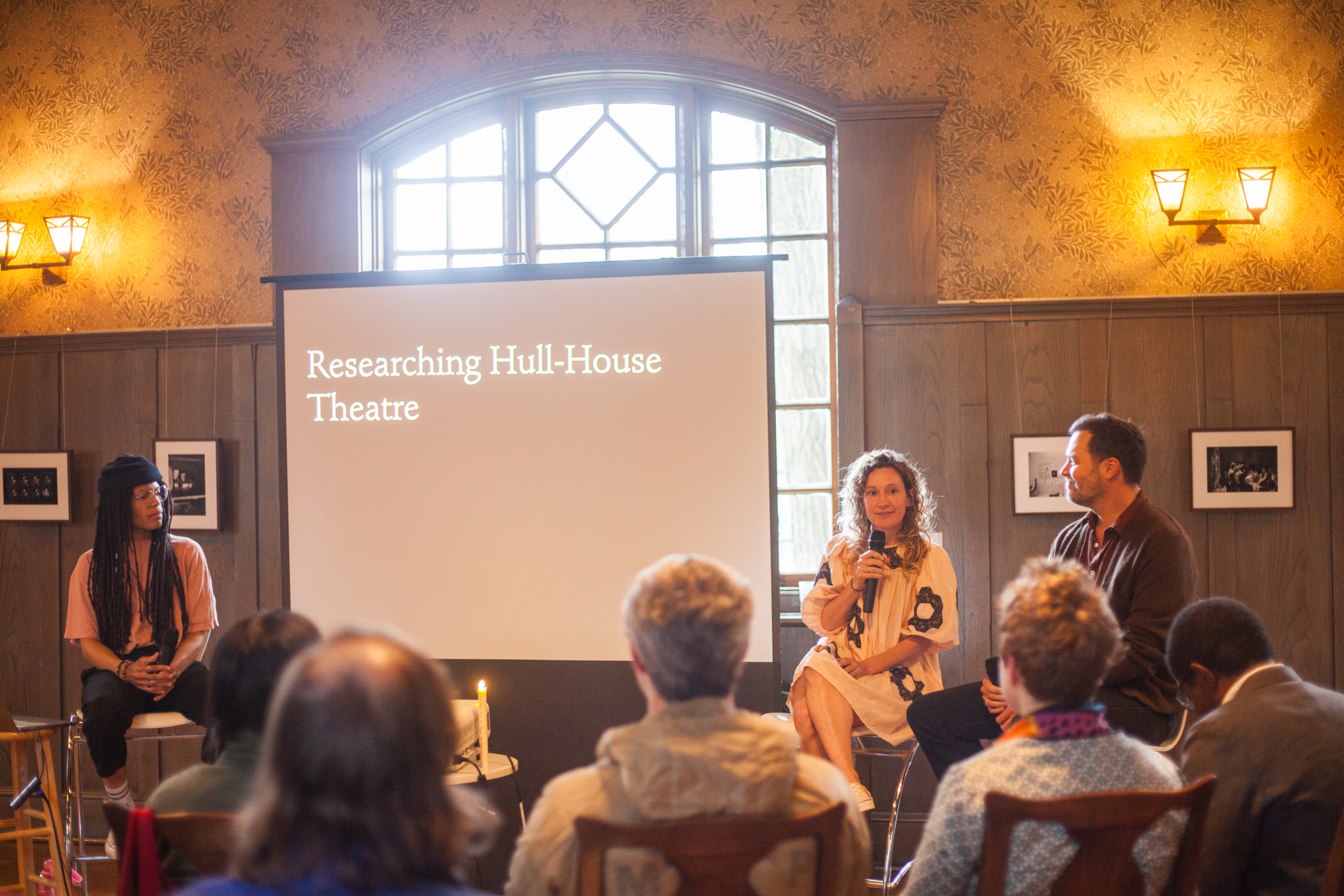 Three people sit at the front of a room with an audience, discussing Researching Hull-House Theatre as shown on a large projector screen, in a warmly lit room with wood paneling and framed photos.