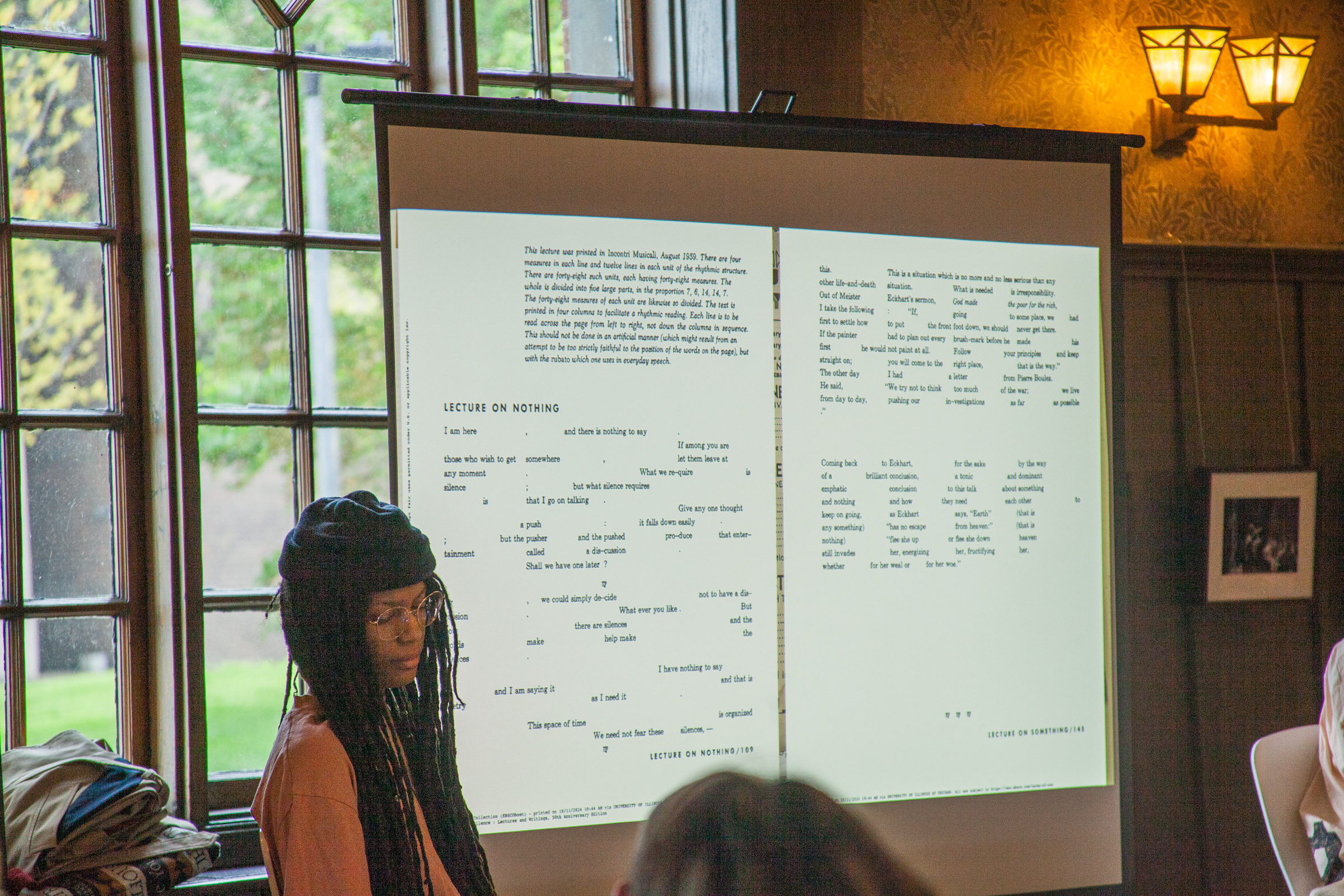 A person with long braided hair and glasses sits near a window beside a projected screen displaying a page of poetry or text in a well-lit room with wood-paneled walls and decorative sconces.