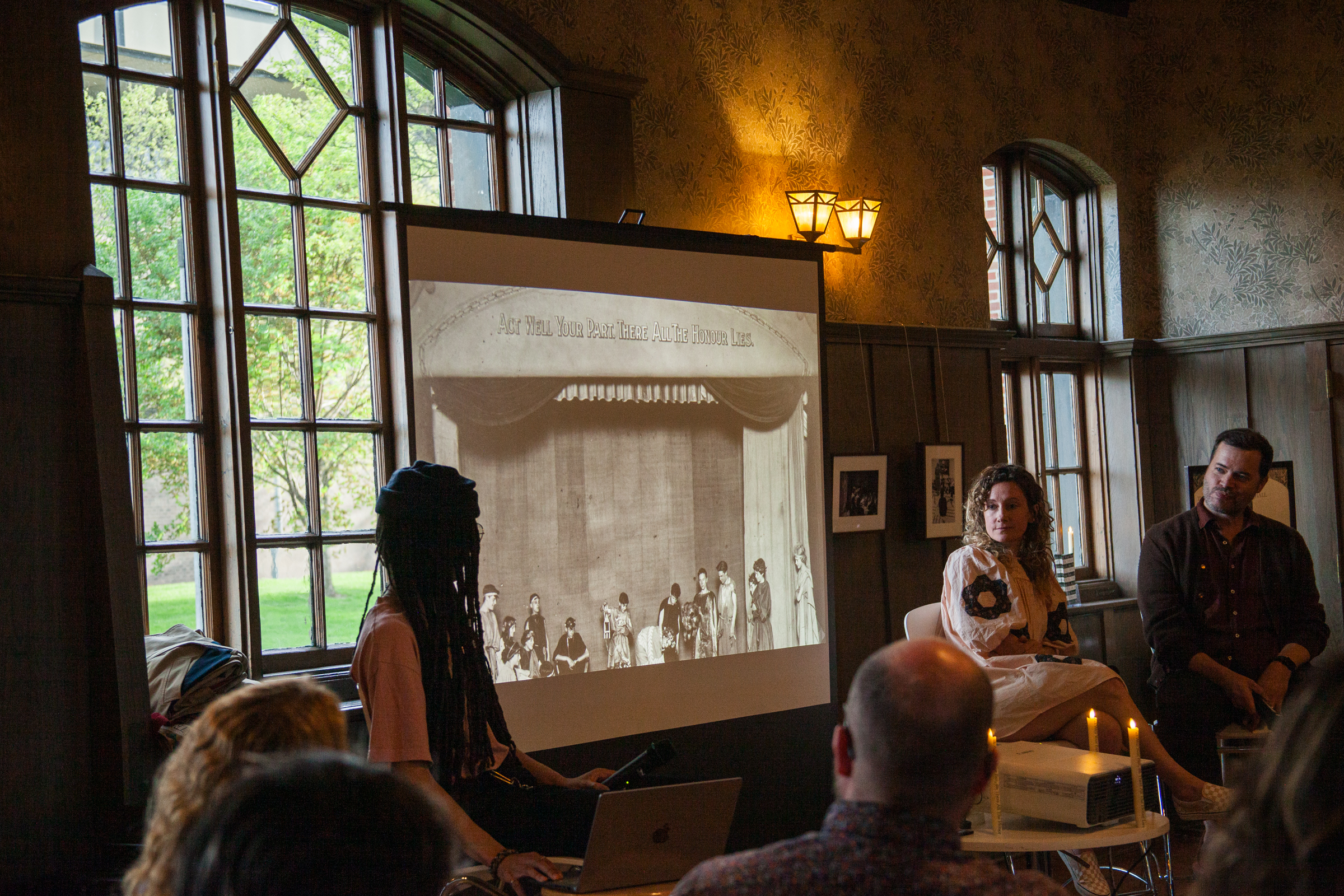 A group of people sit in a dimly lit room watching a black-and-white photo projected onto a screen. The room has large windows, wooden walls, and framed pictures. Some attendees face the presenter.