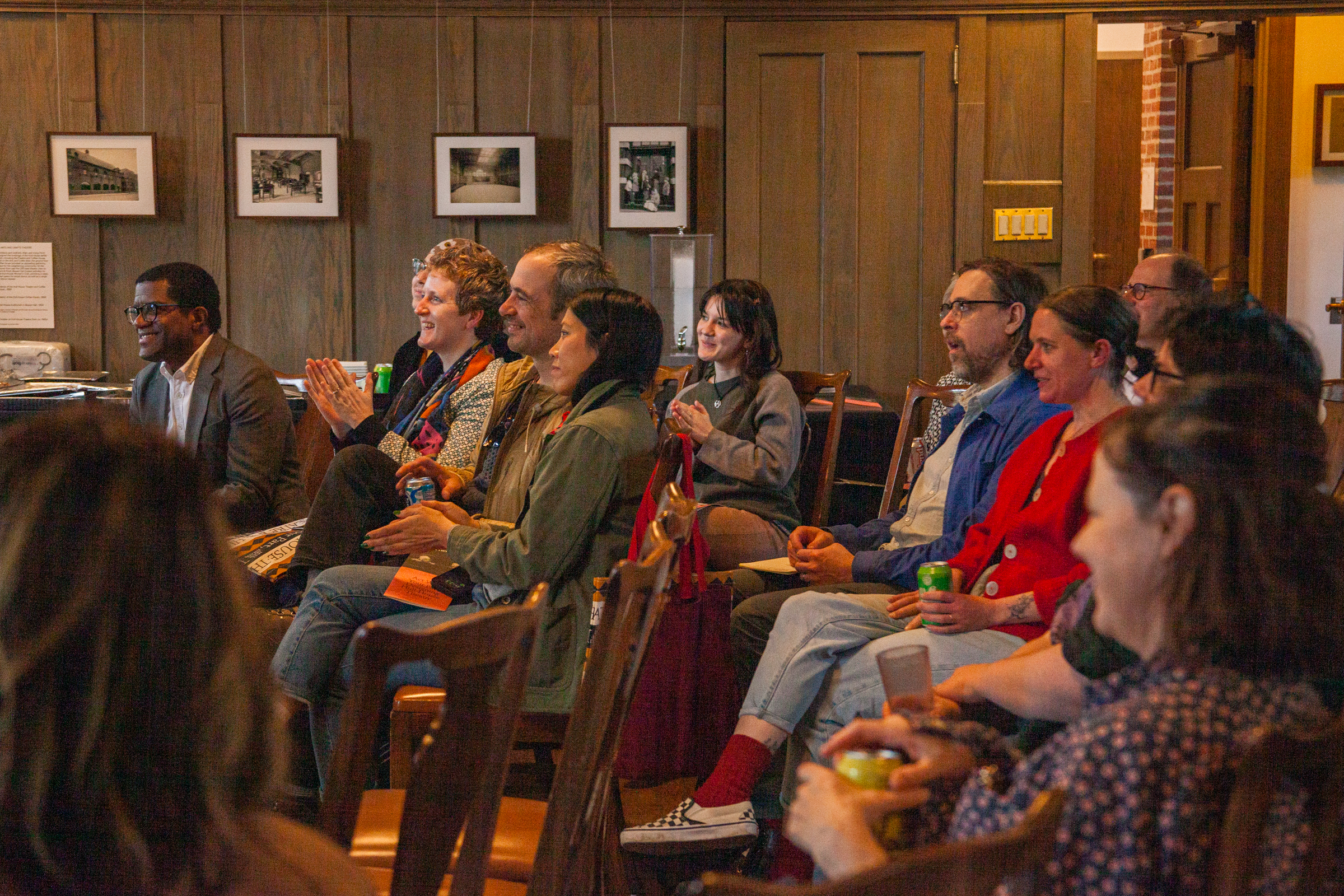 A group of people sit in rows of chairs indoors, smiling and laughing as they watch something off-camera. The room has wood-paneled walls and framed photos. The atmosphere appears warm and lively.
