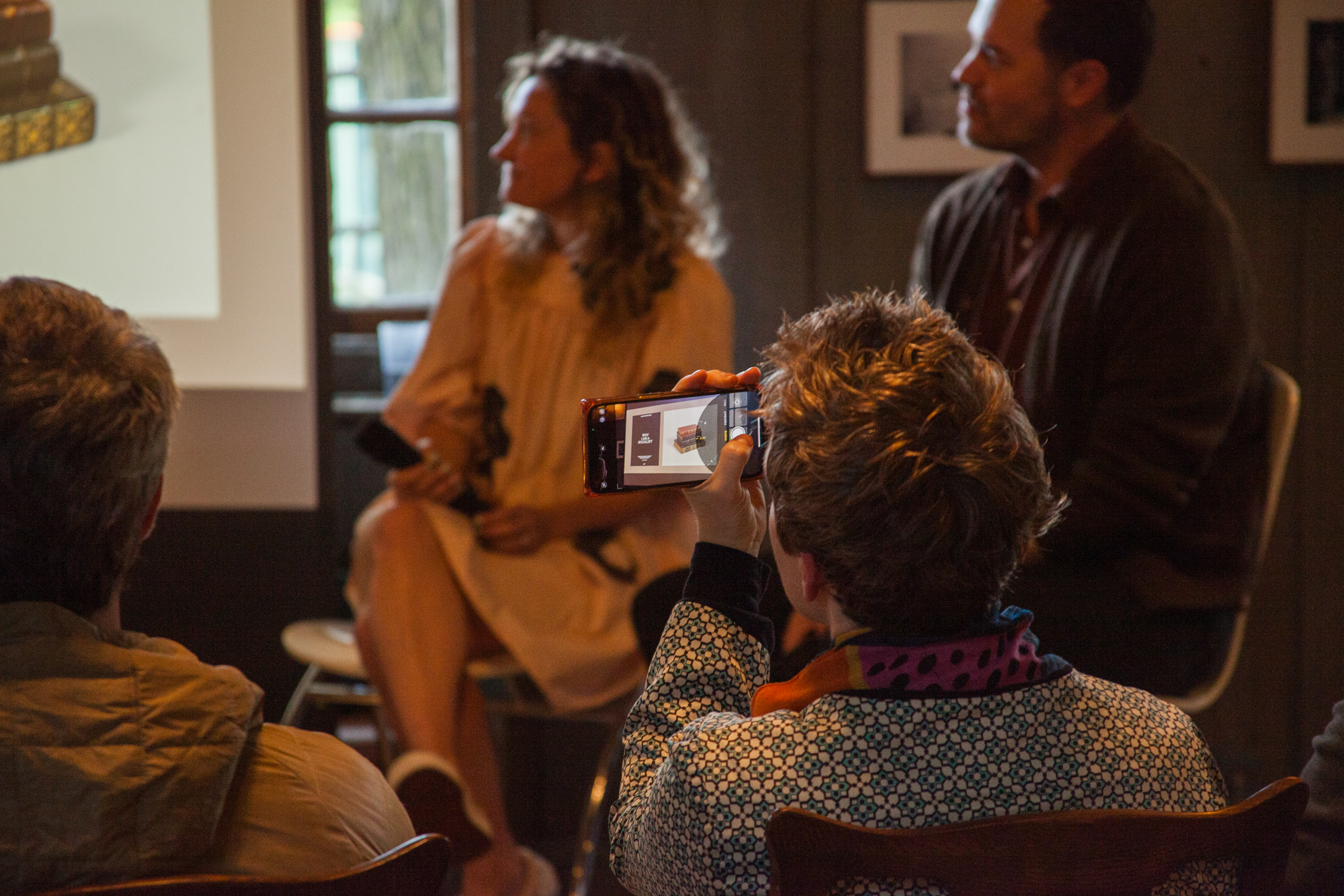 A person in the audience holds up a phone to take a photo of a woman and man seated at the front of a dimly lit room during a presentation or discussion.