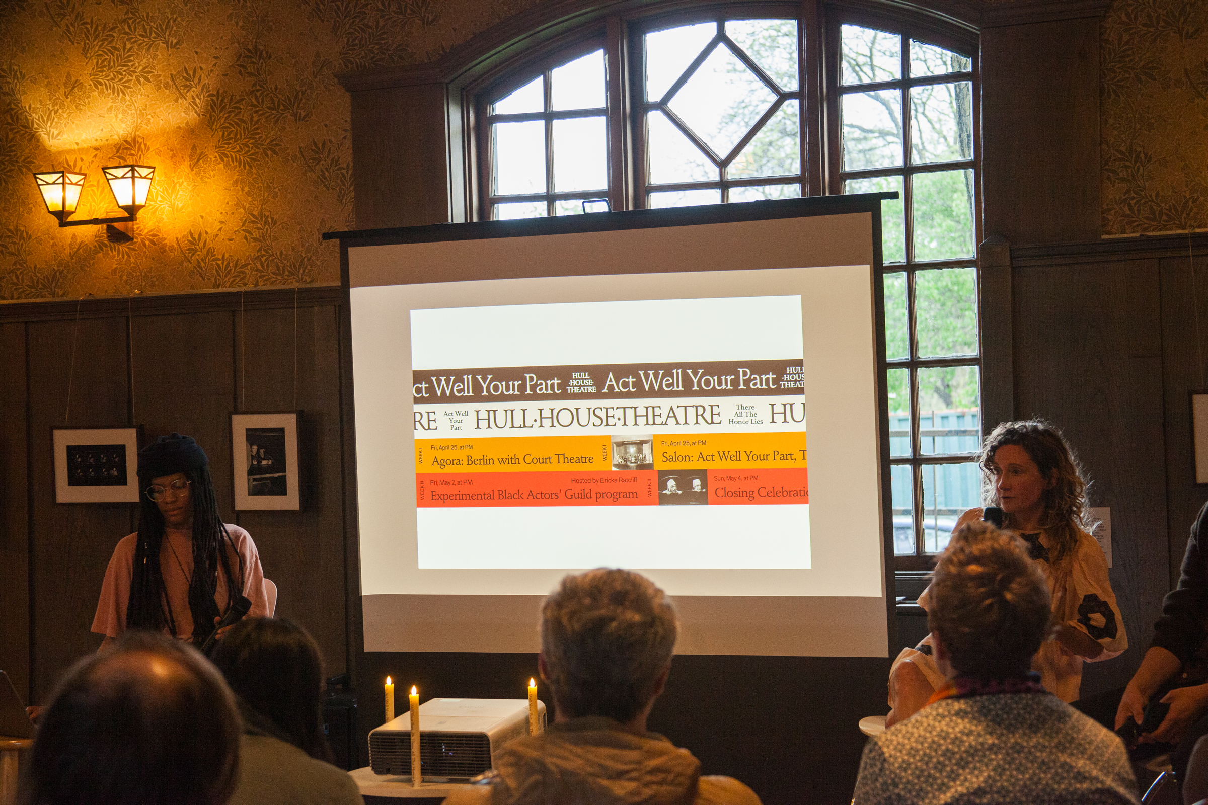 A group of people sit facing a screen in a dimly lit room with wooden walls. The screen displays a presentation titled Act Well Your Part: HULL-HOUSE THEATRE. Two presenters stand at the front near lit candles.